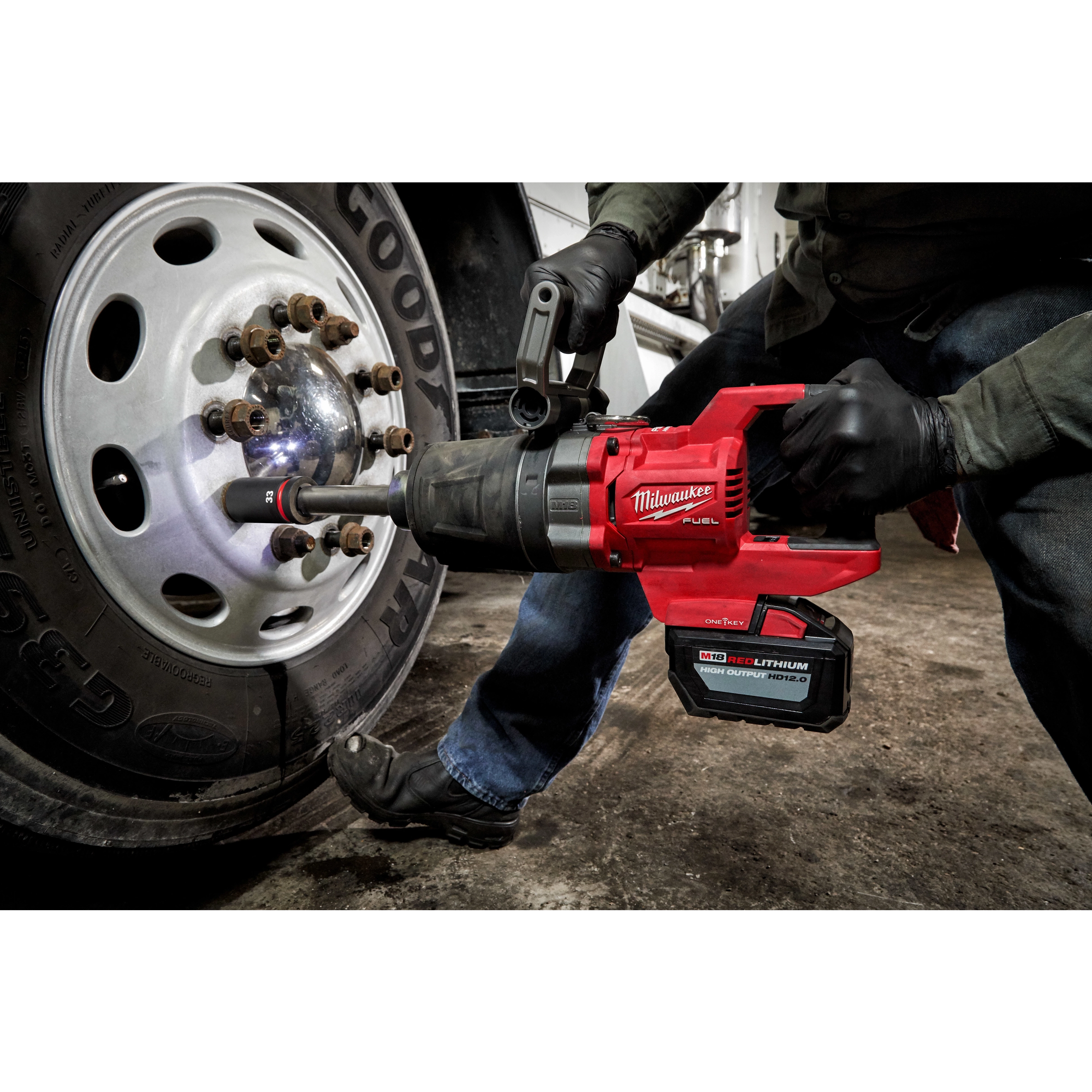 A person uses the M18 FUEL™ 1" D-Handle Extended Anvil High Torque Impact Wrench w/ONE-KEY™ to remove lug nuts from a truck wheel. The red, battery-powered wrench is in operation, showing sparks. The user wears gloves and the environment appears to be a garage or industrial setting.