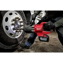A person uses the M18 FUEL™ 1" D-Handle Extended Anvil High Torque Impact Wrench w/ONE-KEY™ to remove lug nuts from a truck wheel. The red, battery-powered wrench is in operation, showing sparks. The user wears gloves and the environment appears to be a garage or industrial setting.
