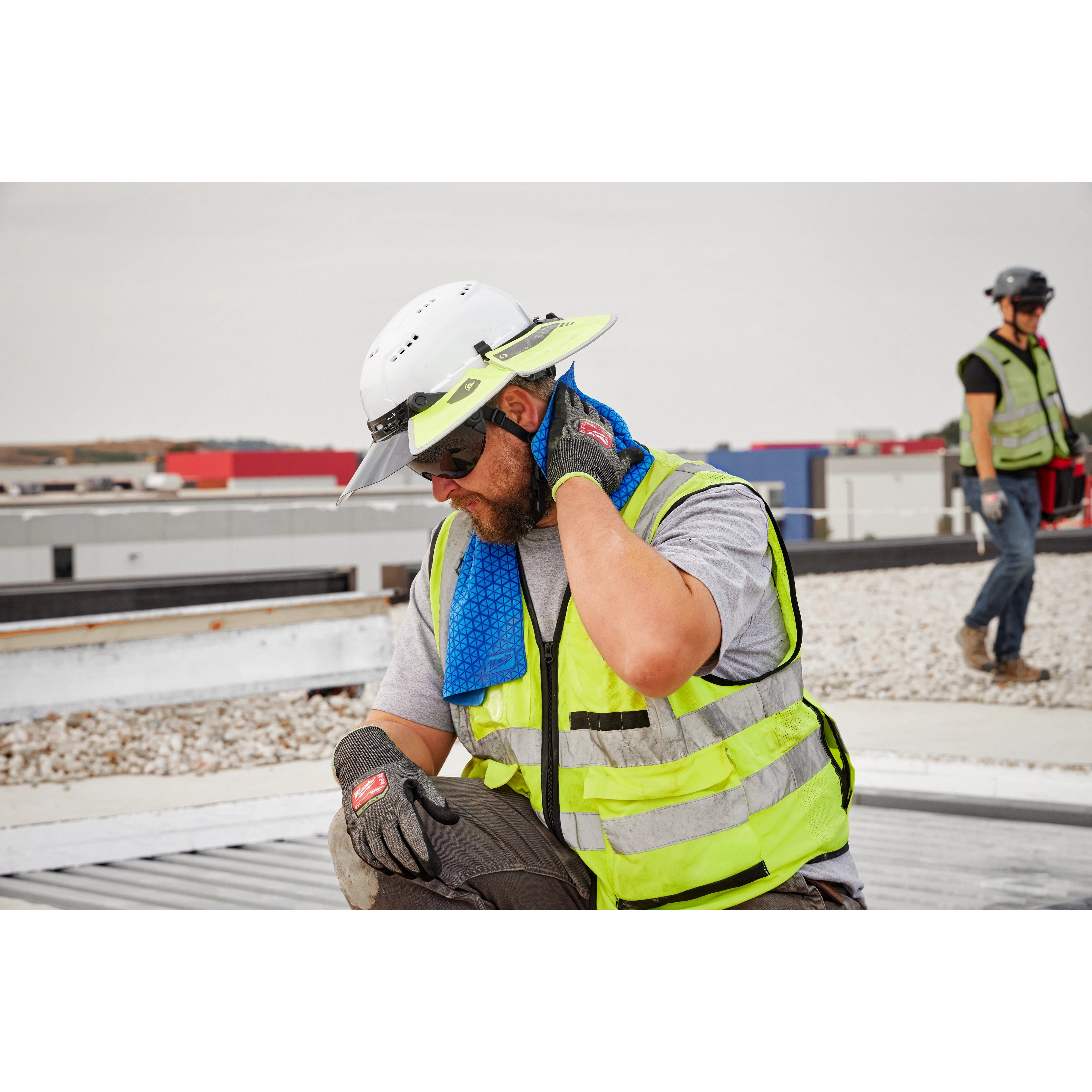 A construction worker, wearing a white hard hat and yellow vest, uses a blue Cooling PVA Towel around their neck on a rooftop. Another worker in a similar outfit is visible in the background.