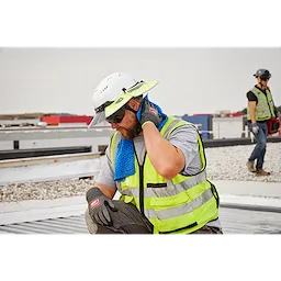 A construction worker, wearing a white hard hat and yellow vest, uses a blue Cooling PVA Towel around their neck on a rooftop. Another worker in a similar outfit is visible in the background.