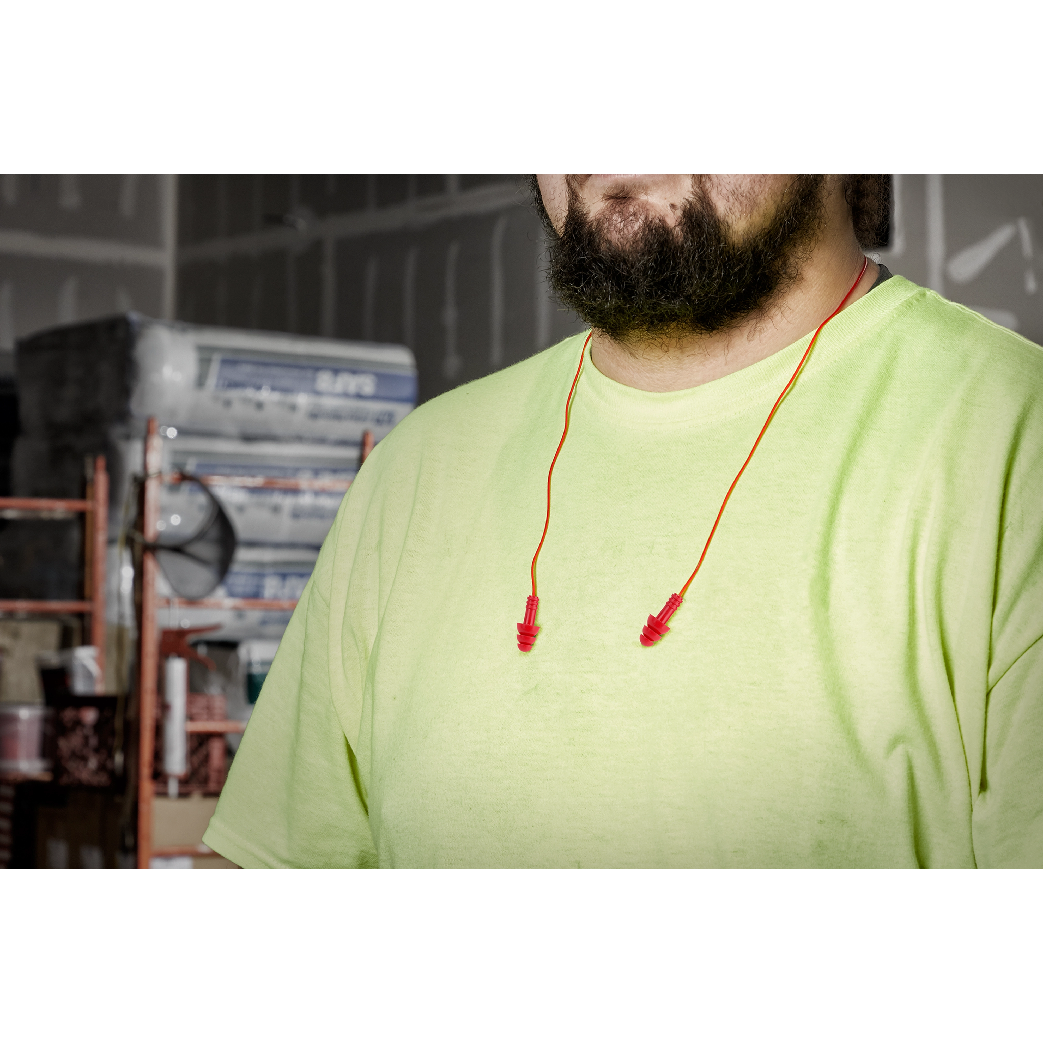 A man with a beard and a neon green shirt wears red Ear Plugs around his neck while standing in a construction setting.