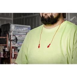 A man with a beard and a neon green shirt wears red Ear Plugs around his neck while standing in a construction setting.