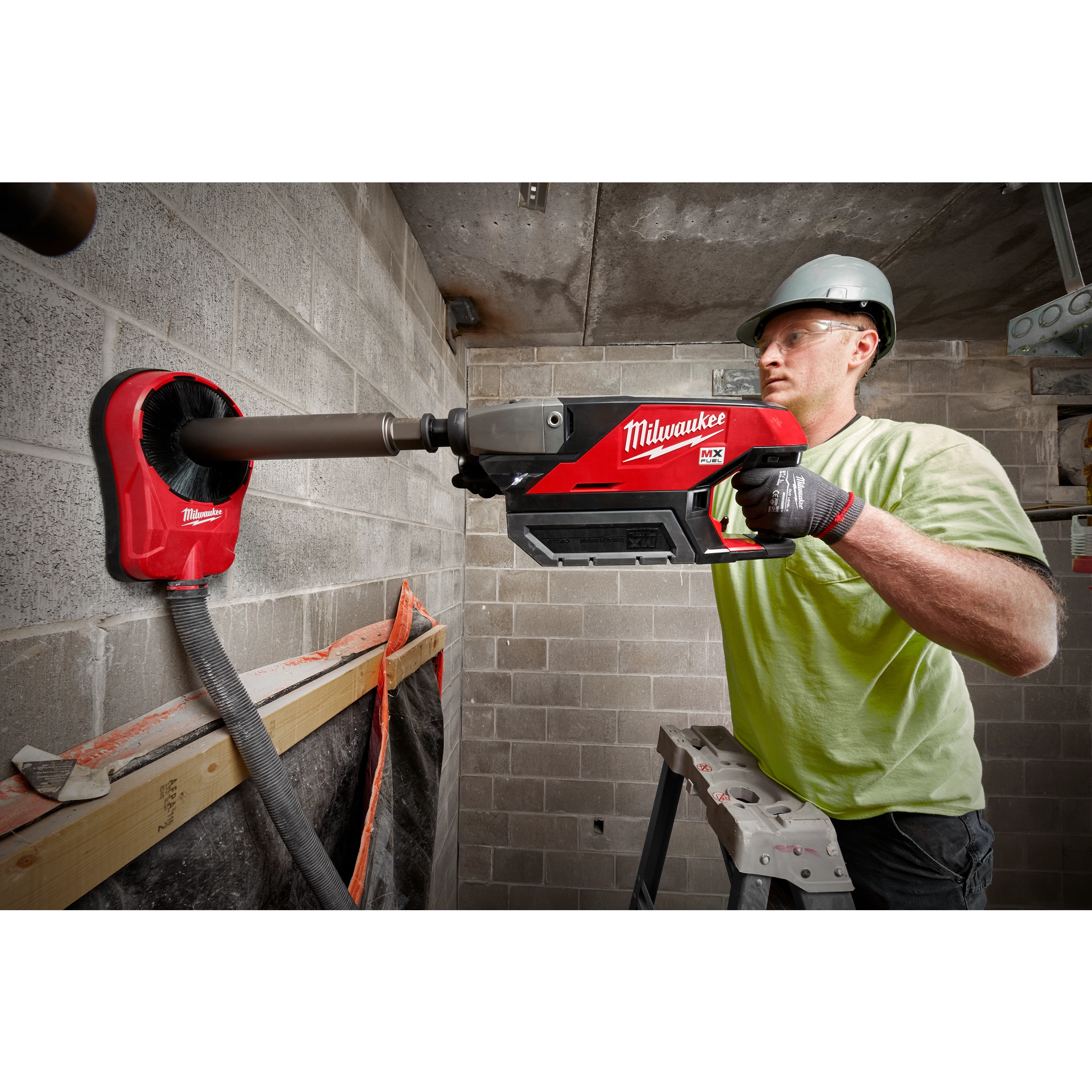 A construction worker operates the Milwaukee MX FUEL™ Handheld Core Drill to create a hole in a concrete wall. The tool is connected to a dust collection system and is red with black and silver accents. The worker is standing on a ladder.