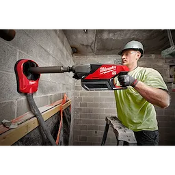 A construction worker operates the Milwaukee MX FUEL™ Handheld Core Drill to create a hole in a concrete wall. The tool is connected to a dust collection system and is red with black and silver accents. The worker is standing on a ladder.