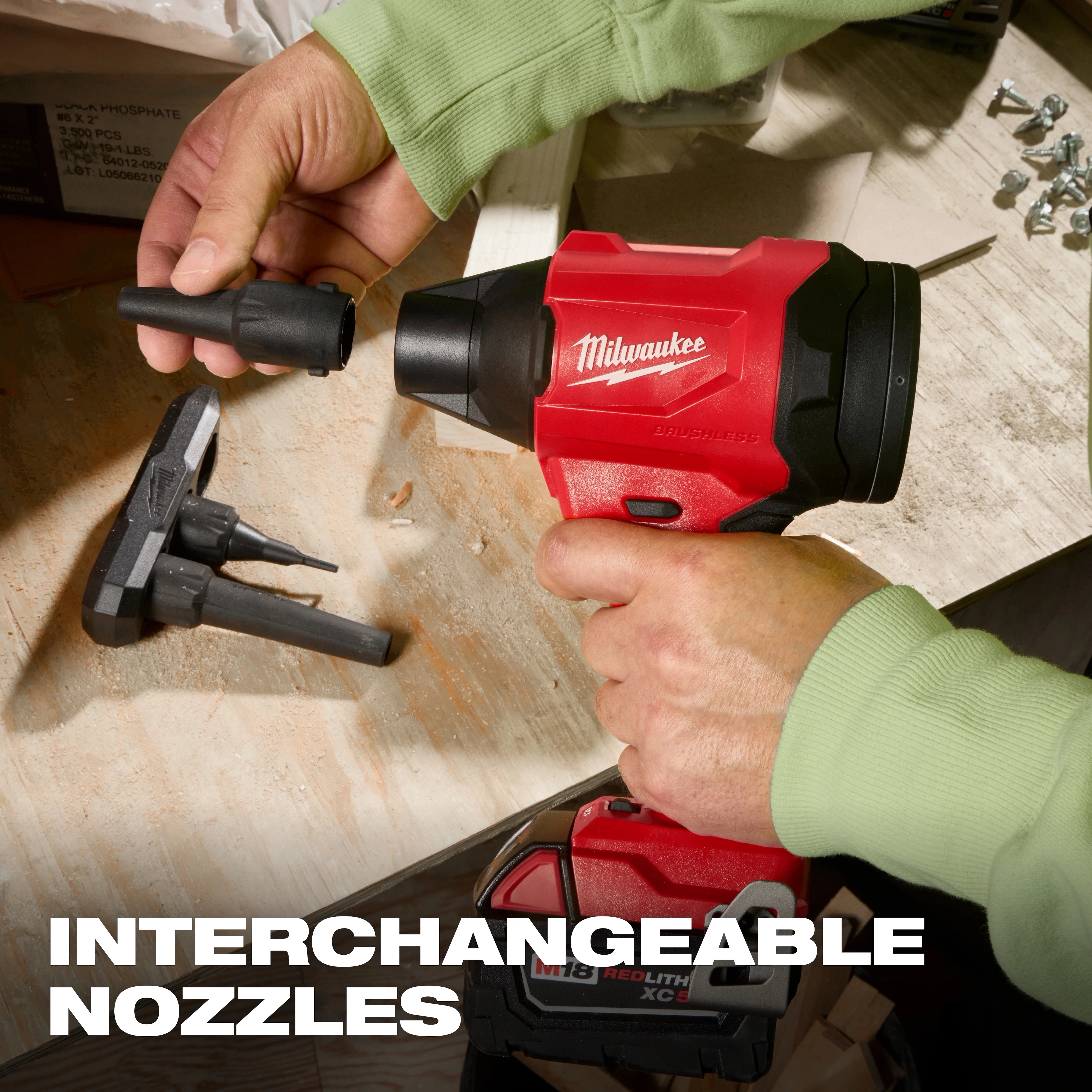 The M18™ Brushless Precision Blower by Milwaukee is shown with interchangeable nozzles. A person in green sleeves is attaching one of the nozzles to the red and black blower. Spare nozzles are placed on the workbench, and the "INTERCHANGEABLE NOZZLES" text is displayed at the bottom.