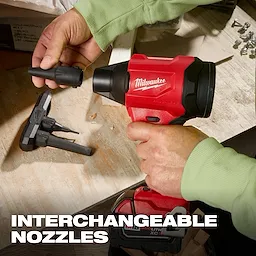 The M18™ Brushless Precision Blower by Milwaukee is shown with interchangeable nozzles. A person in green sleeves is attaching one of the nozzles to the red and black blower. Spare nozzles are placed on the workbench, and the "INTERCHANGEABLE NOZZLES" text is displayed at the bottom.
