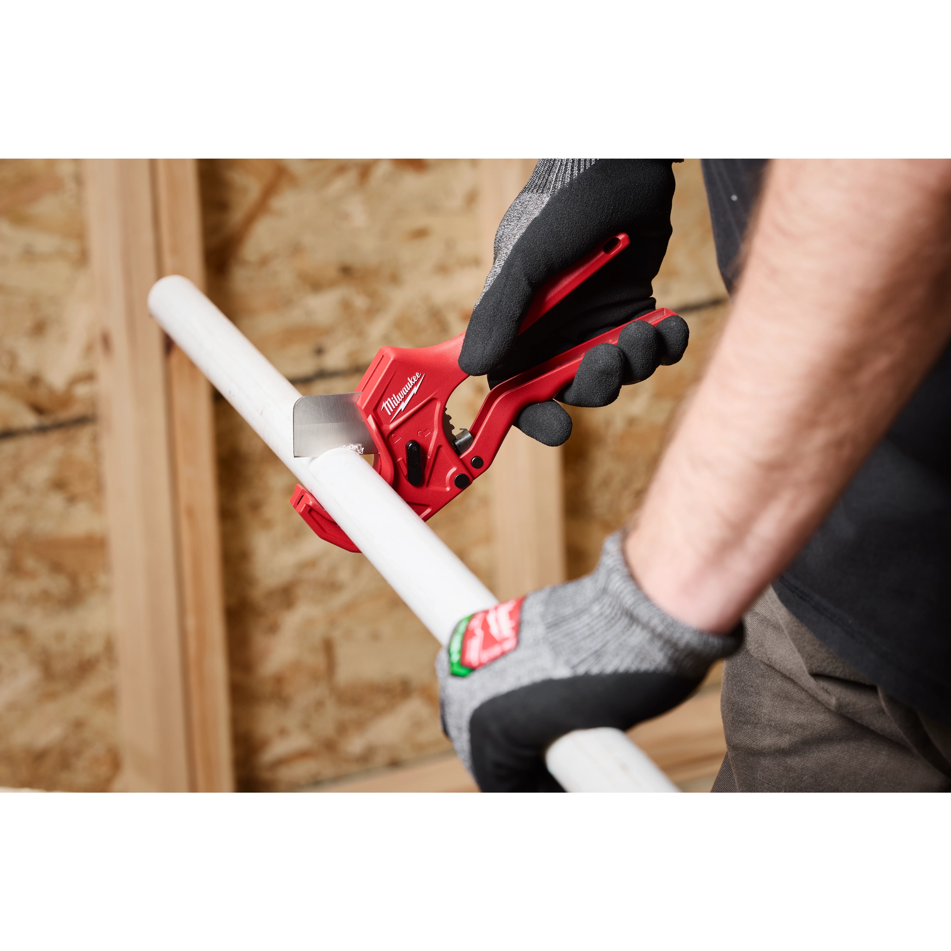 Person using 1-5/8" Ratcheting Pipe Cutter to cut a white PVC pipe, wearing black and gray gloves, with a wood background.