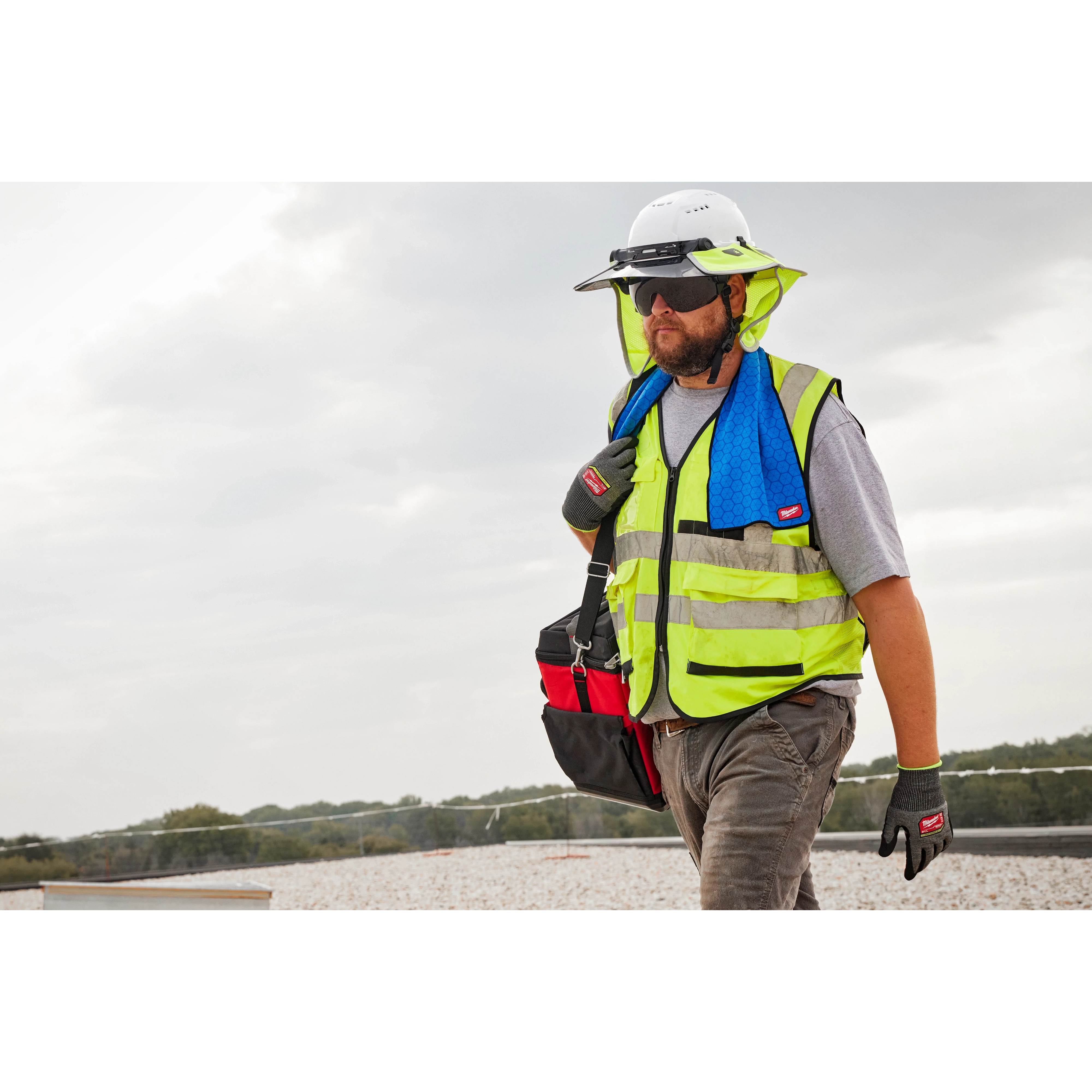 A construction worker wearing a high-visibility vest and a white hard hat carries a bag with a blue Cooling Towel around his neck.