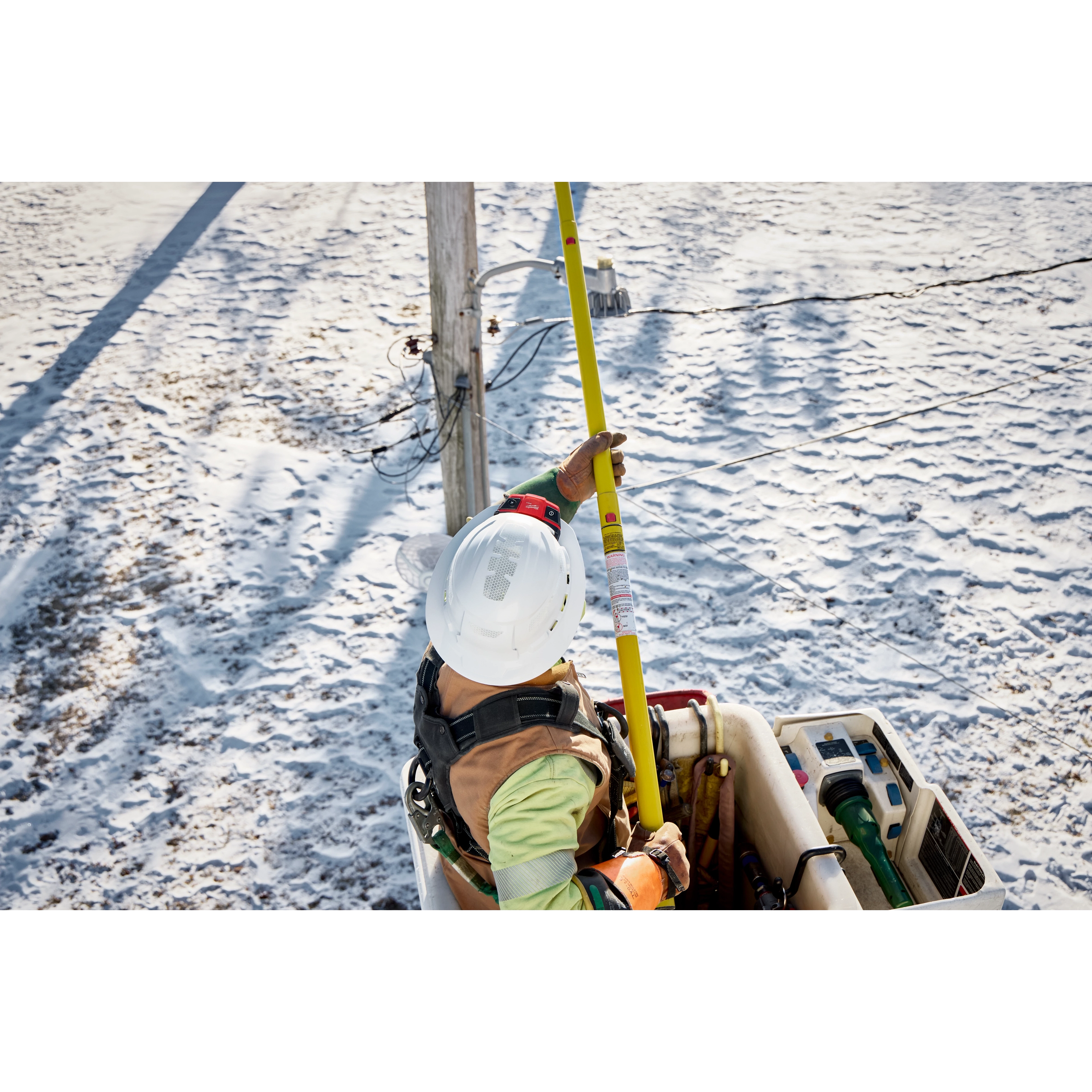 Worker using BOLT Personal Voltage Detector on a yellow pole to check electrical lines from a bucket truck in a snowy area.