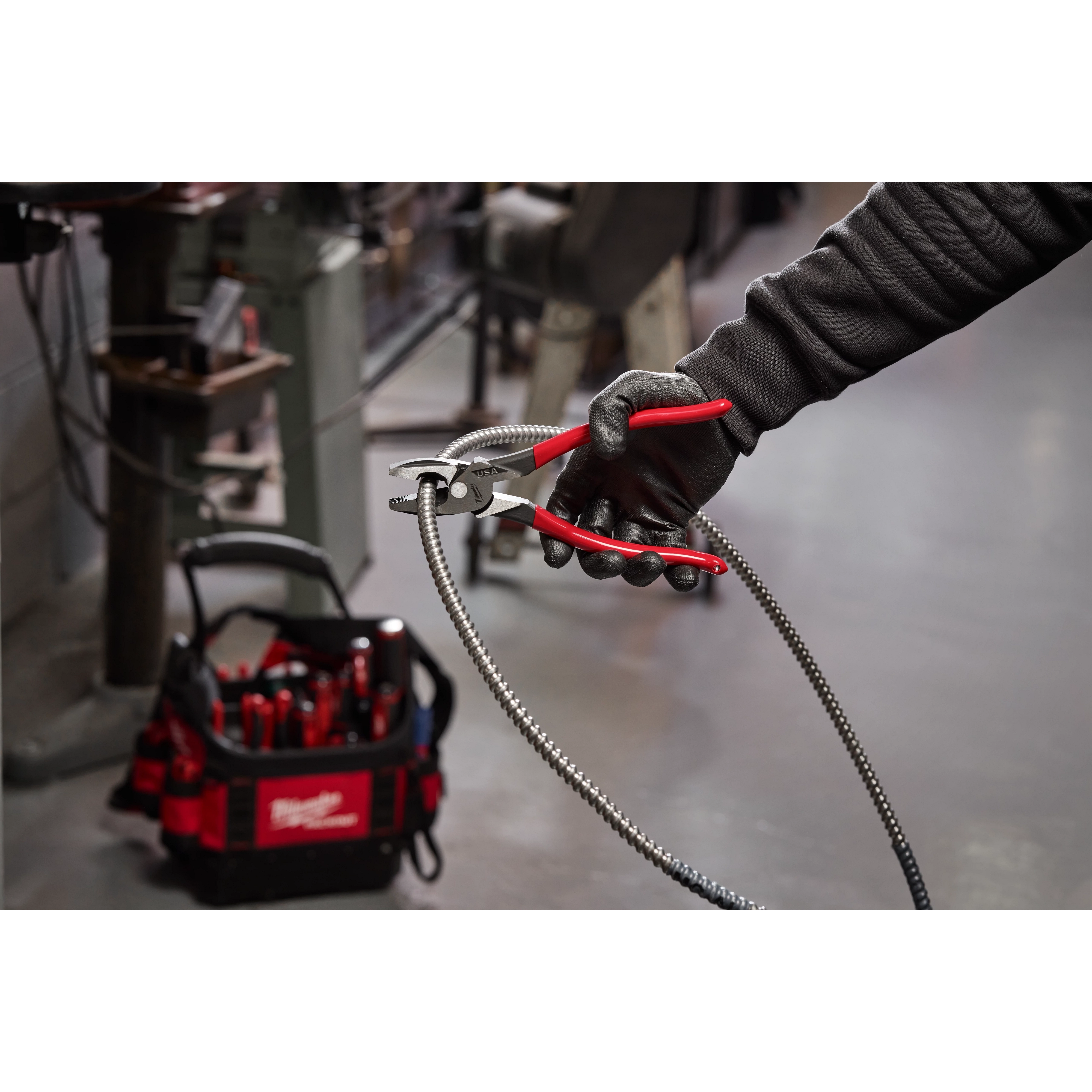 A person wearing a black glove holds red-handled pliers gripping a flexible metal conduit. In the background, a Milwaukee tool bag with various tools is on the floor in a workshop setting.