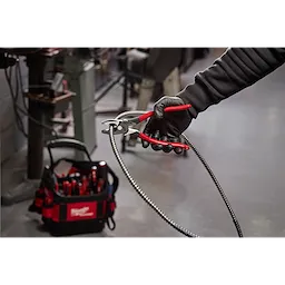 A person wearing a black glove holds red-handled pliers gripping a flexible metal conduit. In the background, a Milwaukee tool bag with various tools is on the floor in a workshop setting.