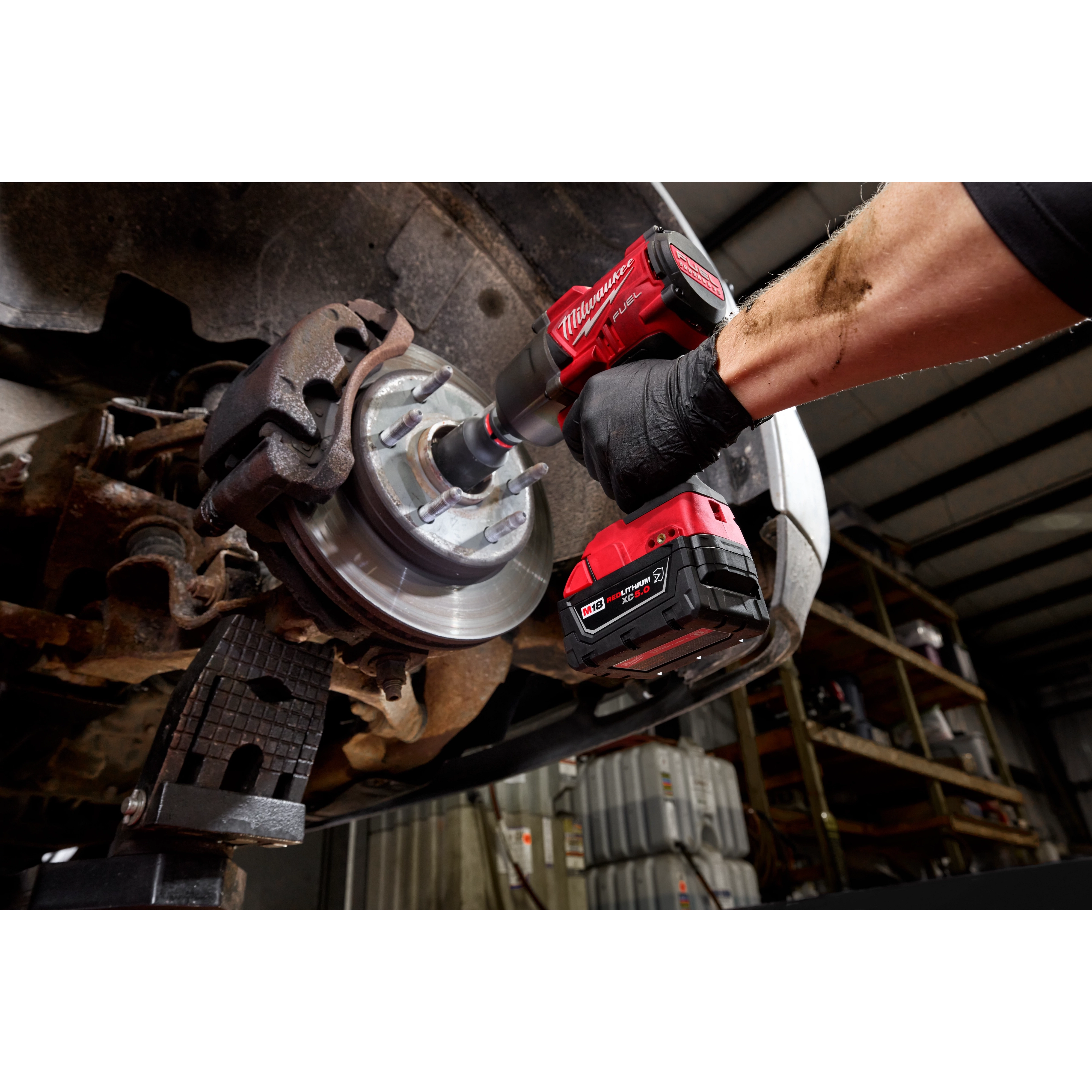 A person is using a Milwaukee M18™ REDLITHIUM™ XC5.0 Resistant Battery attached to a cordless drill to work on a vehicle's brake rotor. The scene is inside a garage, focusing on the heavy-duty application of the battery.