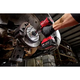 A person is using a Milwaukee M18™ REDLITHIUM™ XC5.0 Resistant Battery attached to a cordless drill to work on a vehicle's brake rotor. The scene is inside a garage, focusing on the heavy-duty application of the battery.