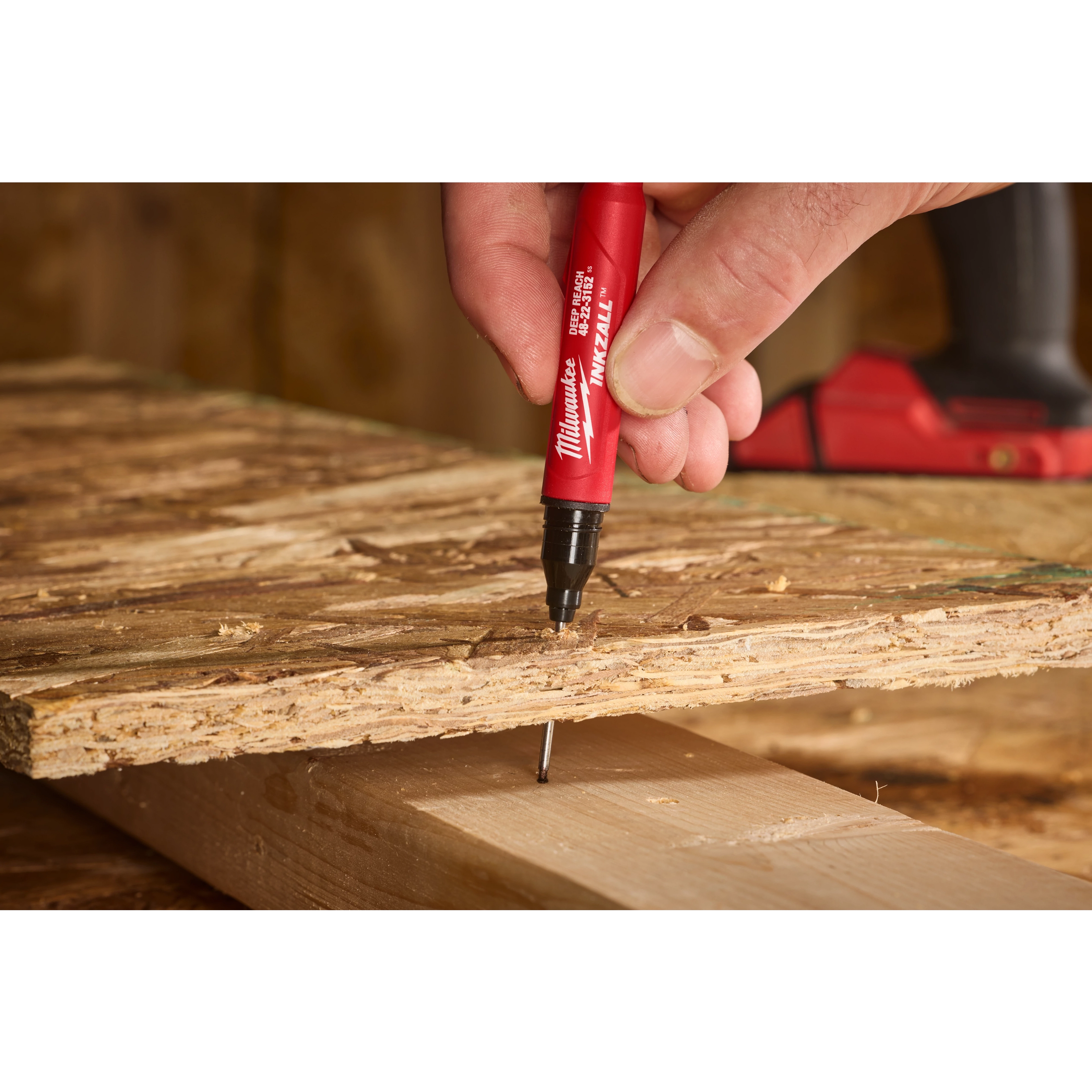 A hand holds the INKZALL™ 2IN1 Deep Reach Ultra Fine Point Black Jobsite Marker, marking a piece of wood with precision. The marker features a red body and a black fine point tip, ideal for detailed work on rough surfaces.