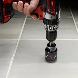 A person uses a red and black drill to cut through gray tiles with a Carbide Grit Hole Saw attached. The hole saw is circular with a grit edge, designed for heavy-duty cutting in hard materials.