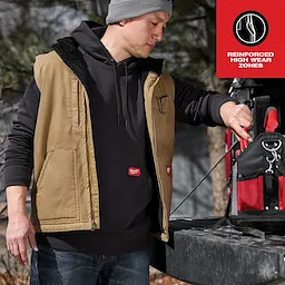 A person wearing a Heavy Duty Sherpa-Lined Vest Feature Shot in khaki color, standing beside a red and black tool bag on a truck.