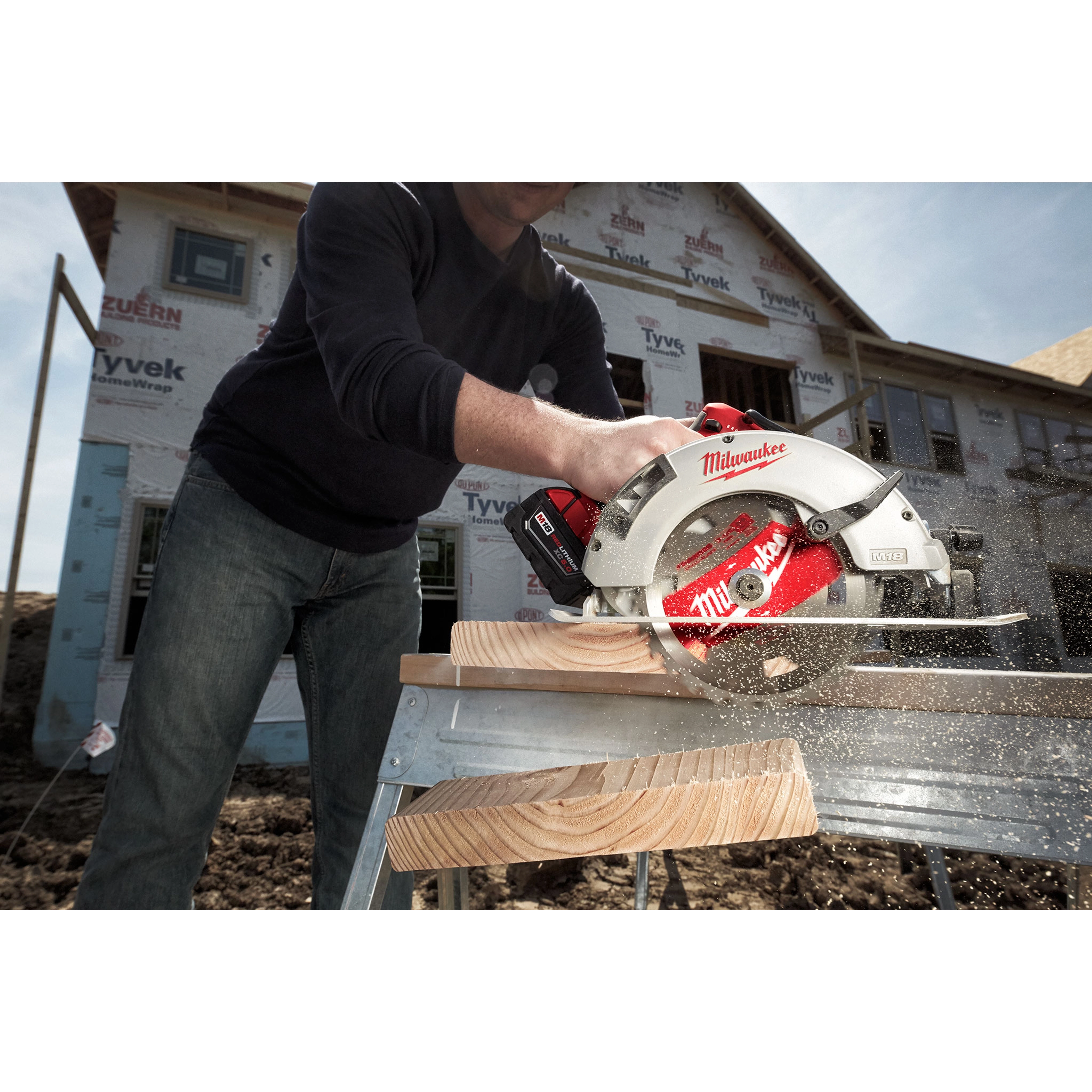 A person uses the Milwaukee 7%201%2F4%22 Brushless Circular Saw to cut through a wooden plank. The saw is red and silver, and the cutting is taking place on a construction site with a partially built house in the background.