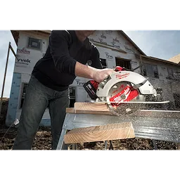 A person uses the Milwaukee 7%201%2F4%22 Brushless Circular Saw to cut through a wooden plank. The saw is red and silver, and the cutting is taking place on a construction site with a partially built house in the background.
