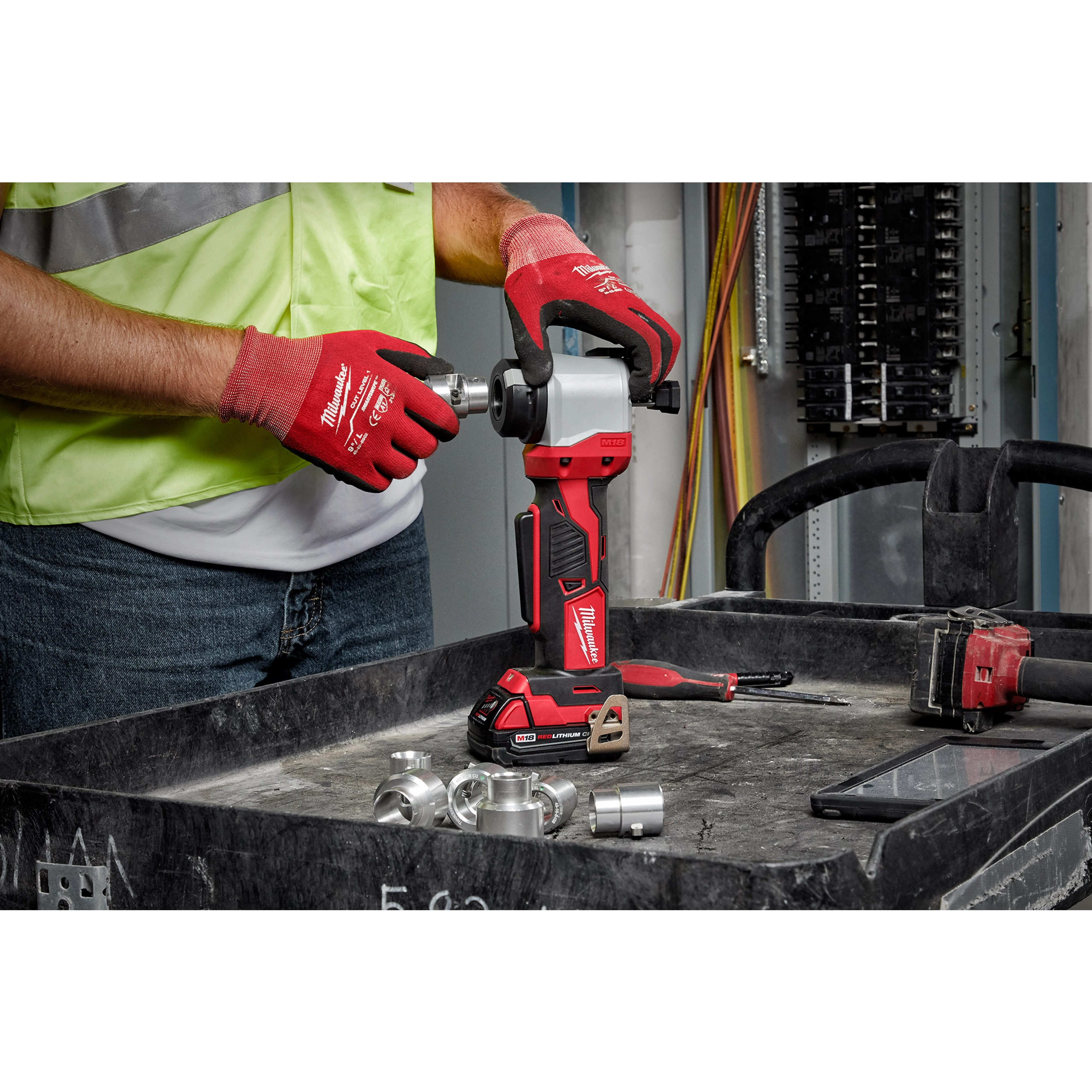 Person using the M18 Cable Stripper in a workshop with gloves, surrounded by metal fittings and tools on a black workbench.