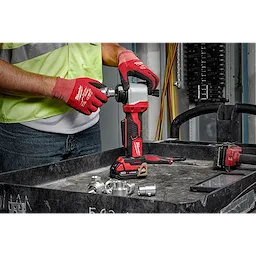 Person using the M18 Cable Stripper in a workshop with gloves, surrounded by metal fittings and tools on a black workbench.