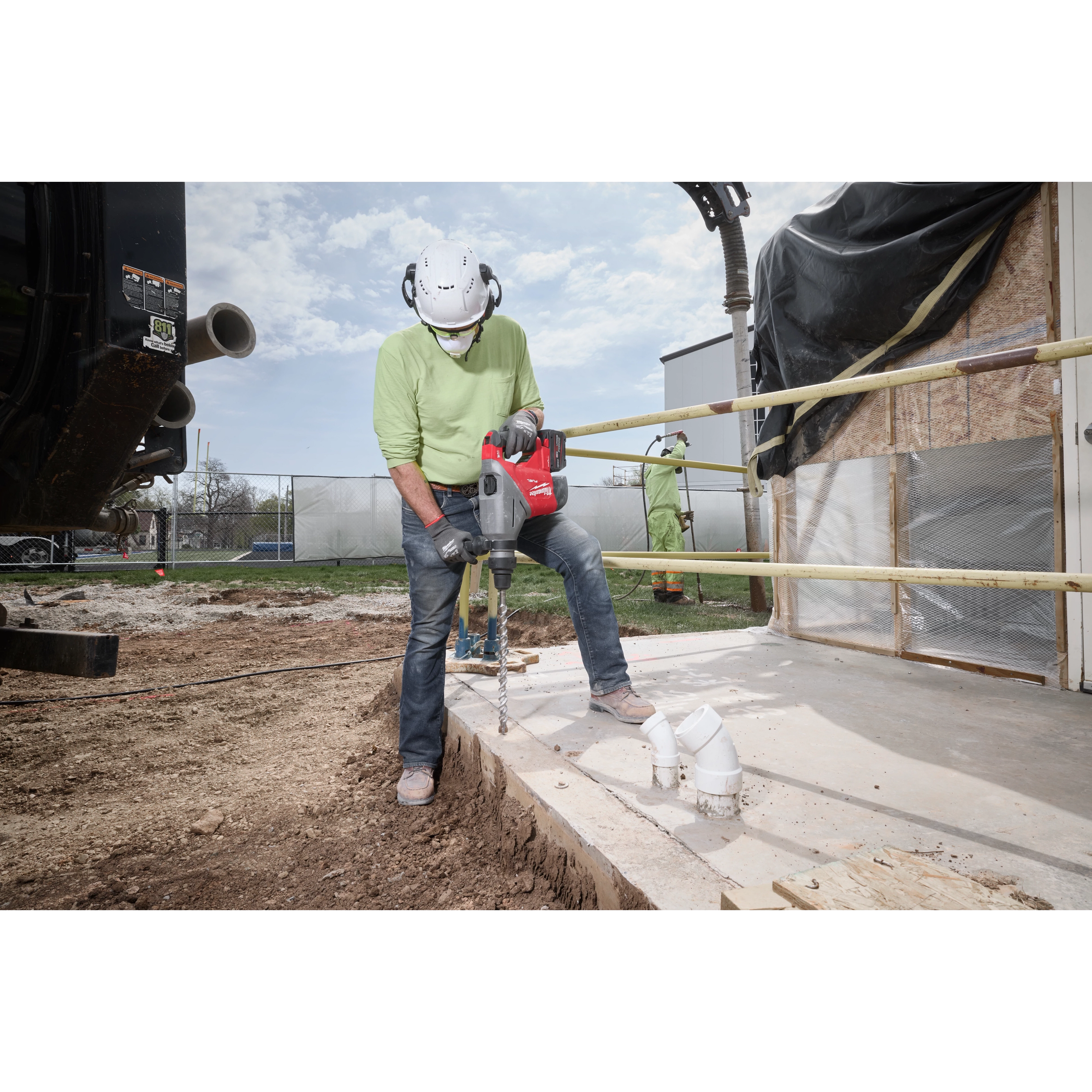 A worker in safety gear uses the M18 FUEL™ 1-3/4" SDS MAX Rotary Hammer Kit w/ ONE-KEY to drill into a concrete surface on a construction site. The powerful tool features enhanced performance technology for heavy-duty drilling tasks. Pipes and construction materials are visible nearby.