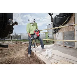 A worker in safety gear uses the M18 FUEL™ 1-3/4" SDS MAX Rotary Hammer Kit w/ ONE-KEY to drill into a concrete surface on a construction site. The powerful tool features enhanced performance technology for heavy-duty drilling tasks. Pipes and construction materials are visible nearby.