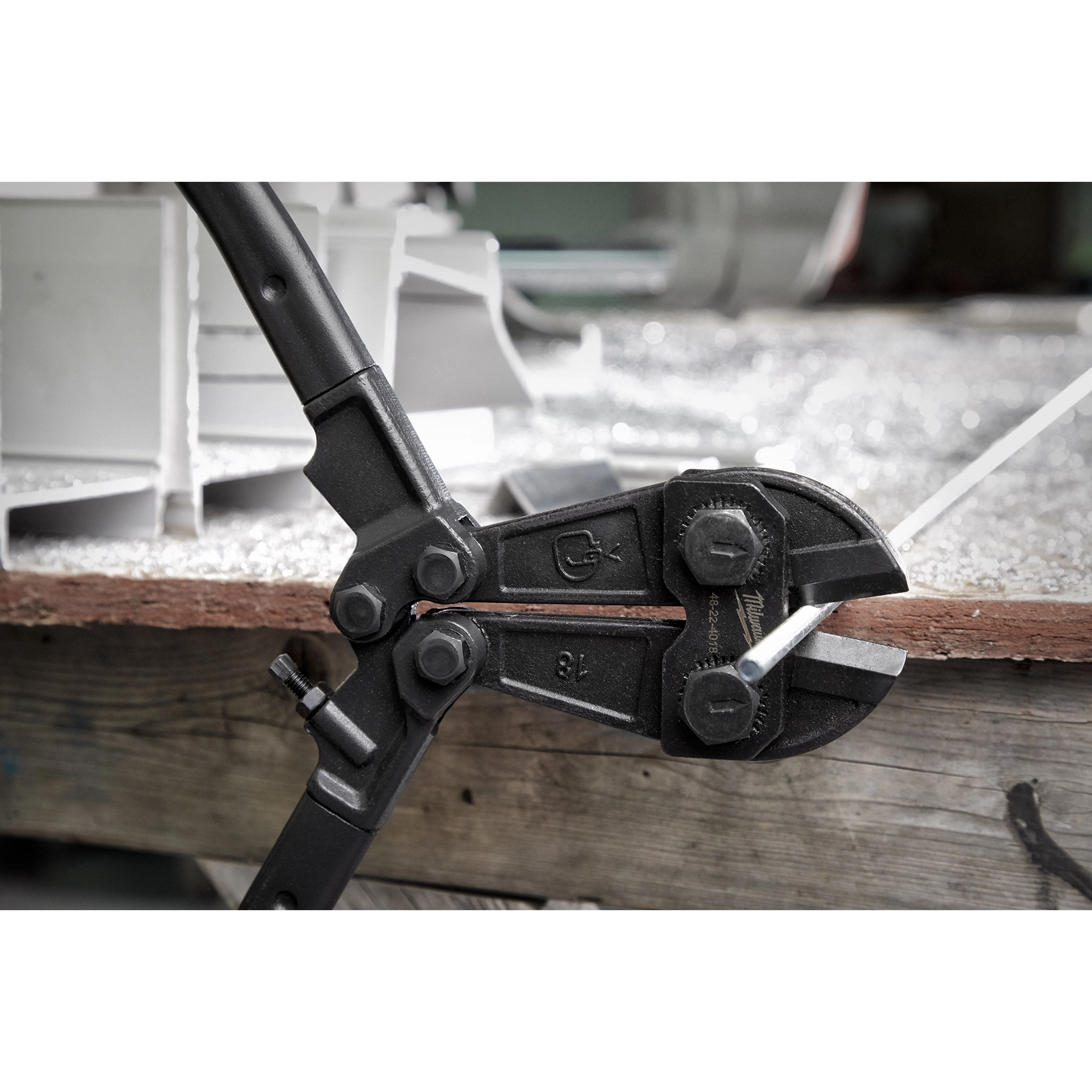 18" Bolt Cutter with black handles cutting a metal rod on a wooden workbench. The tool features two large bolts near the jaw.
