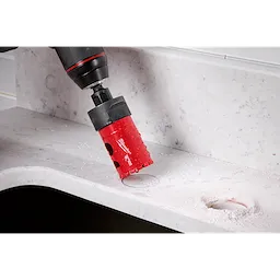 A Milwaukee® 1-3/8" DIAMOND MAX™ Hole Saw attached to a drill is seen cutting through a light gray, stone-like surface. Dust and residue from the cutting are visible around the freshly made hole. Both the tool and the work surface are clearly displayed.