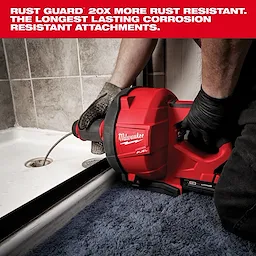 A Milwaukee Fuel drain cleaning tool is being used to unclog a bathroom drain. The red machine is shown in close-up with a worker's gloved hands operating it. Text at the top highlights the Rust Guard feature, stating it is 20 times more rust-resistant.