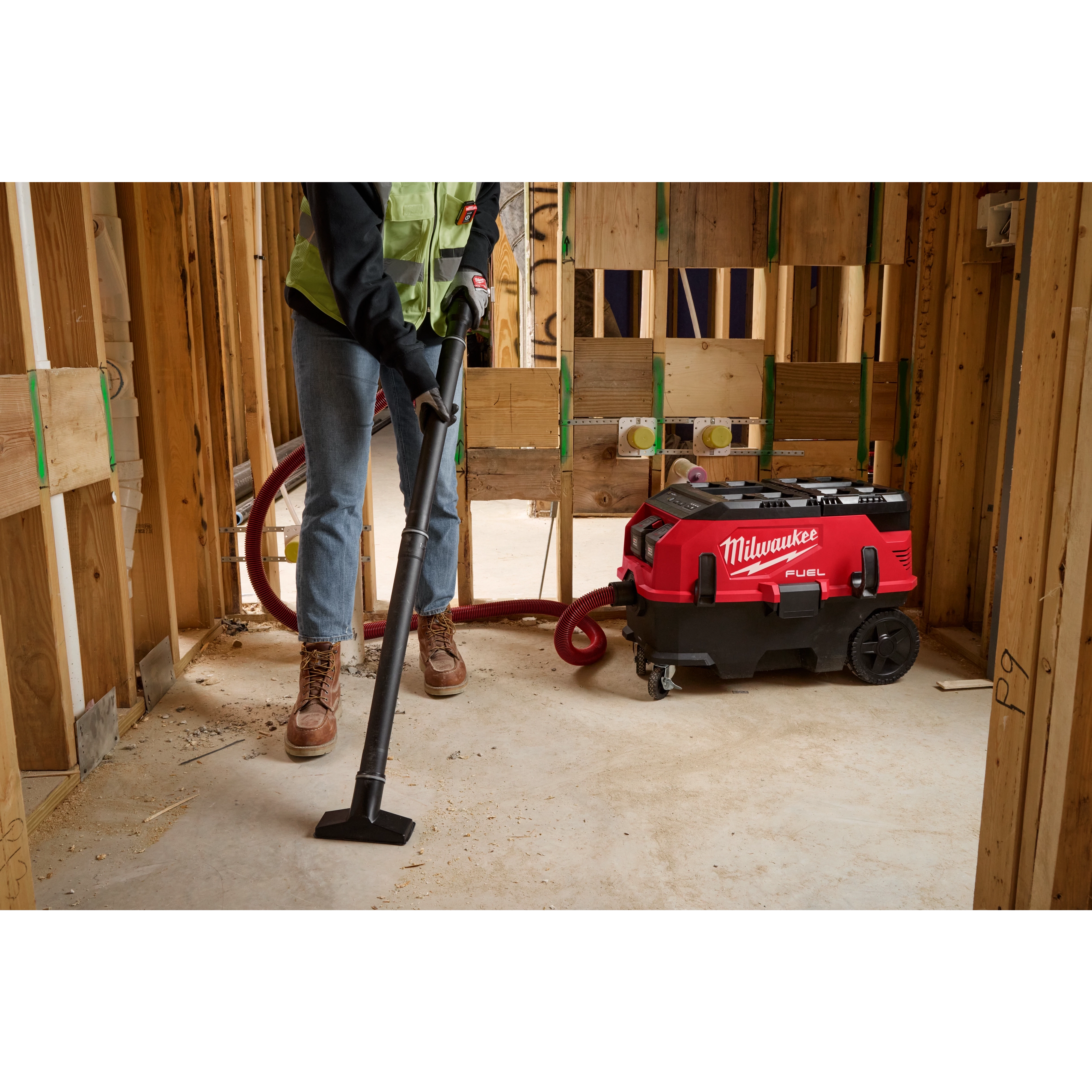 A person is using a vacuum from the PACKOUT™ Cleaning Accessory Kit to clean up a construction site. The vacuum is red and black and has "Milwaukee" written on it. The area is surrounded by unfinished wooden walls.