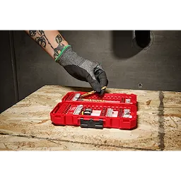 A person wearing gloves is picking a drill bit from an open red case of the SHOCKWAVE™ Impact Duty Drill & Drive Set – 50PC, which is placed on a wooden surface.
