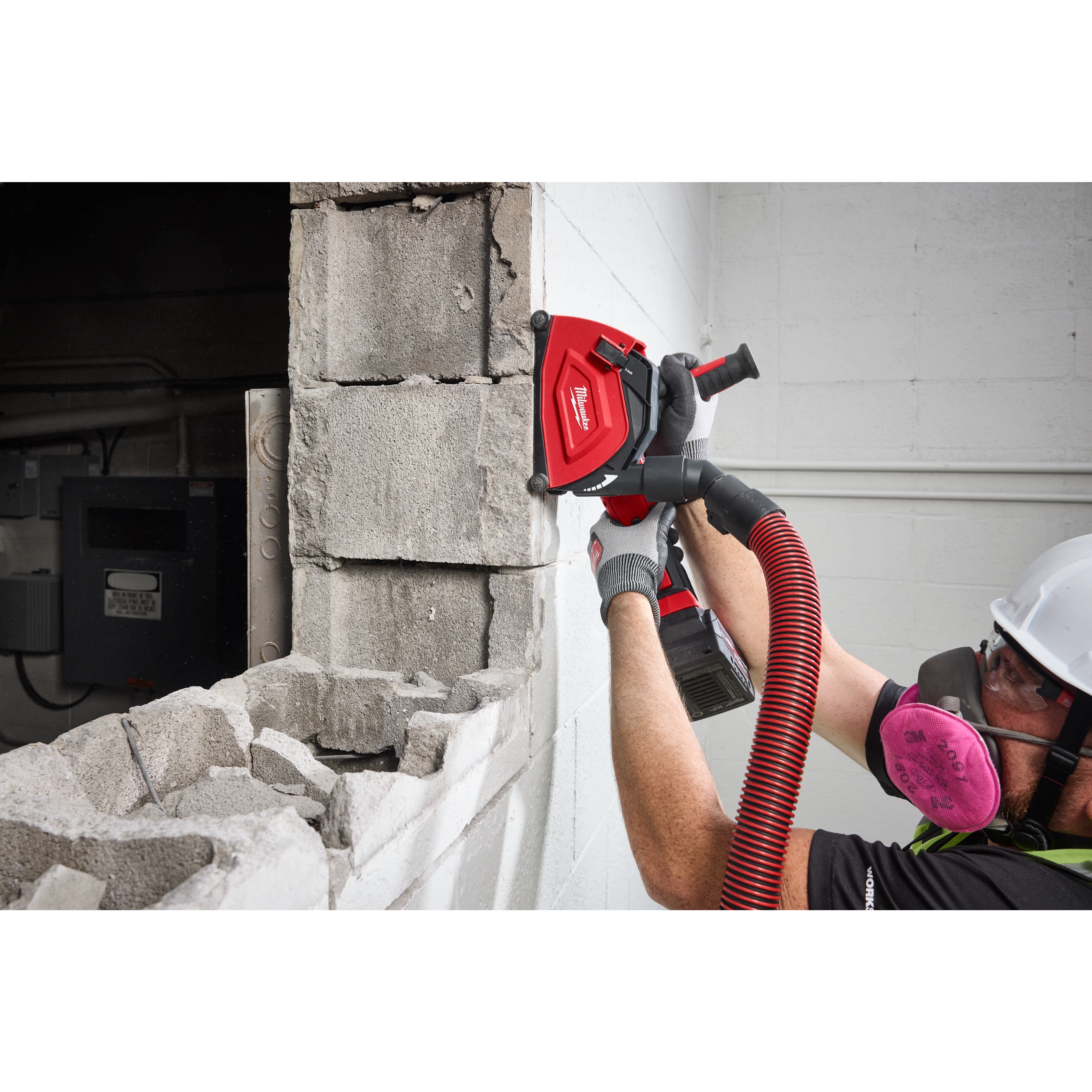 A worker uses a Milwaukee 5" Cutting Dust Shroud attached to a power tool to cut a concrete wall. The shroud is visible, connected to a hose for dust extraction.