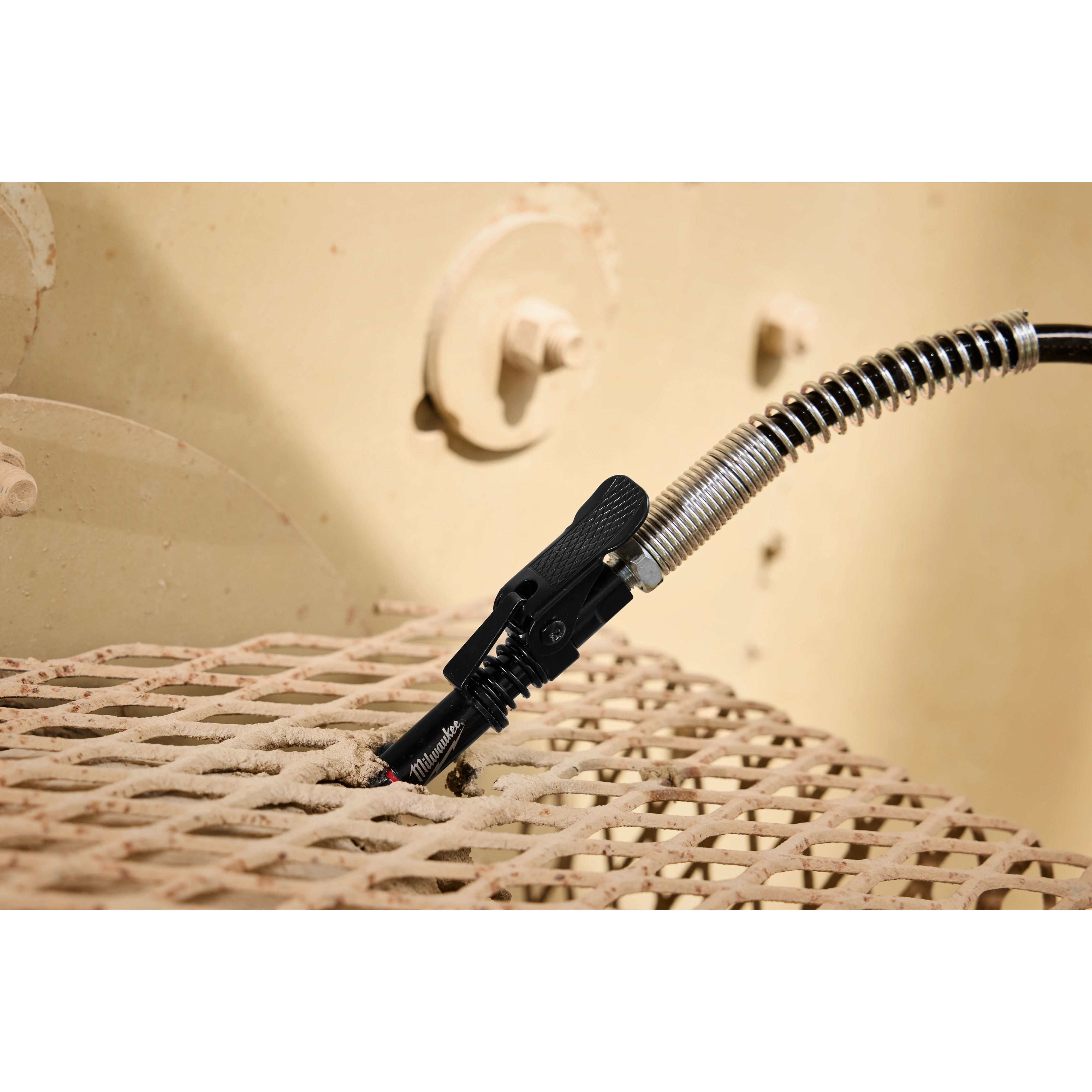 M18 FUEL Grease Gun nozzle and hose positioned on a metal grid surface.