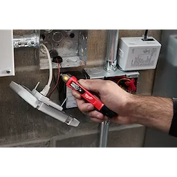 A person is using a Dual Range Voltage Detector w/ LED to check electrical connections in an unfinished metal junction box. The detector has a red and black design with an LED lighted tip indicating voltage presence.