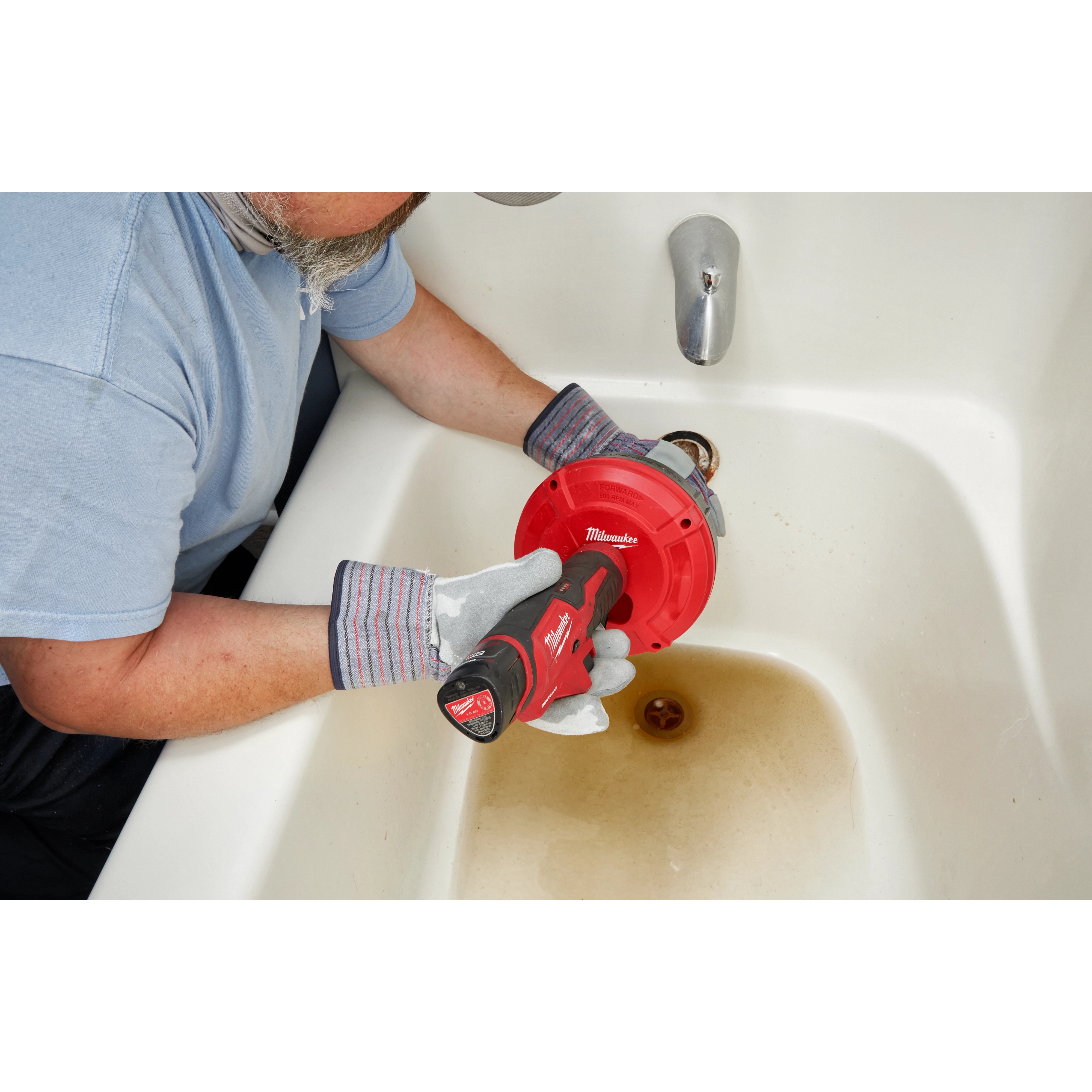 A person uses a TRAPSNAKE 25' Auger in a bathtub drain. The auger is red with a black handle featuring the Milwaukee logo.