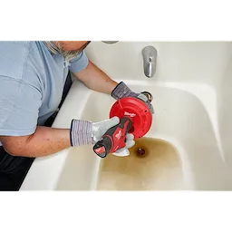 A person uses a TRAPSNAKE 25' Auger in a bathtub drain. The auger is red with a black handle featuring the Milwaukee logo.