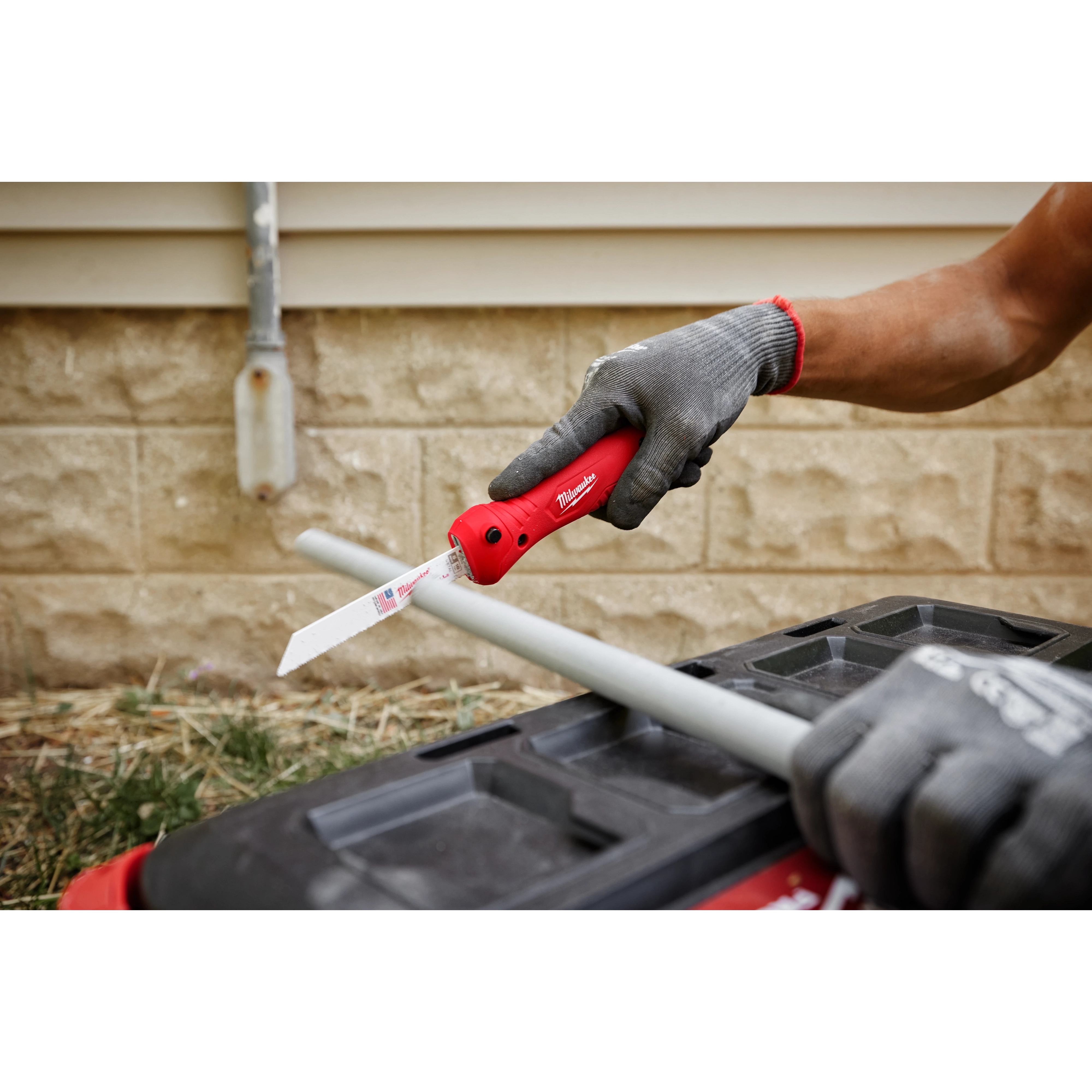 A person wearing grey gloves uses a red Folding Jab Saw to cut a grey pipe against a black surface near a brick wall.