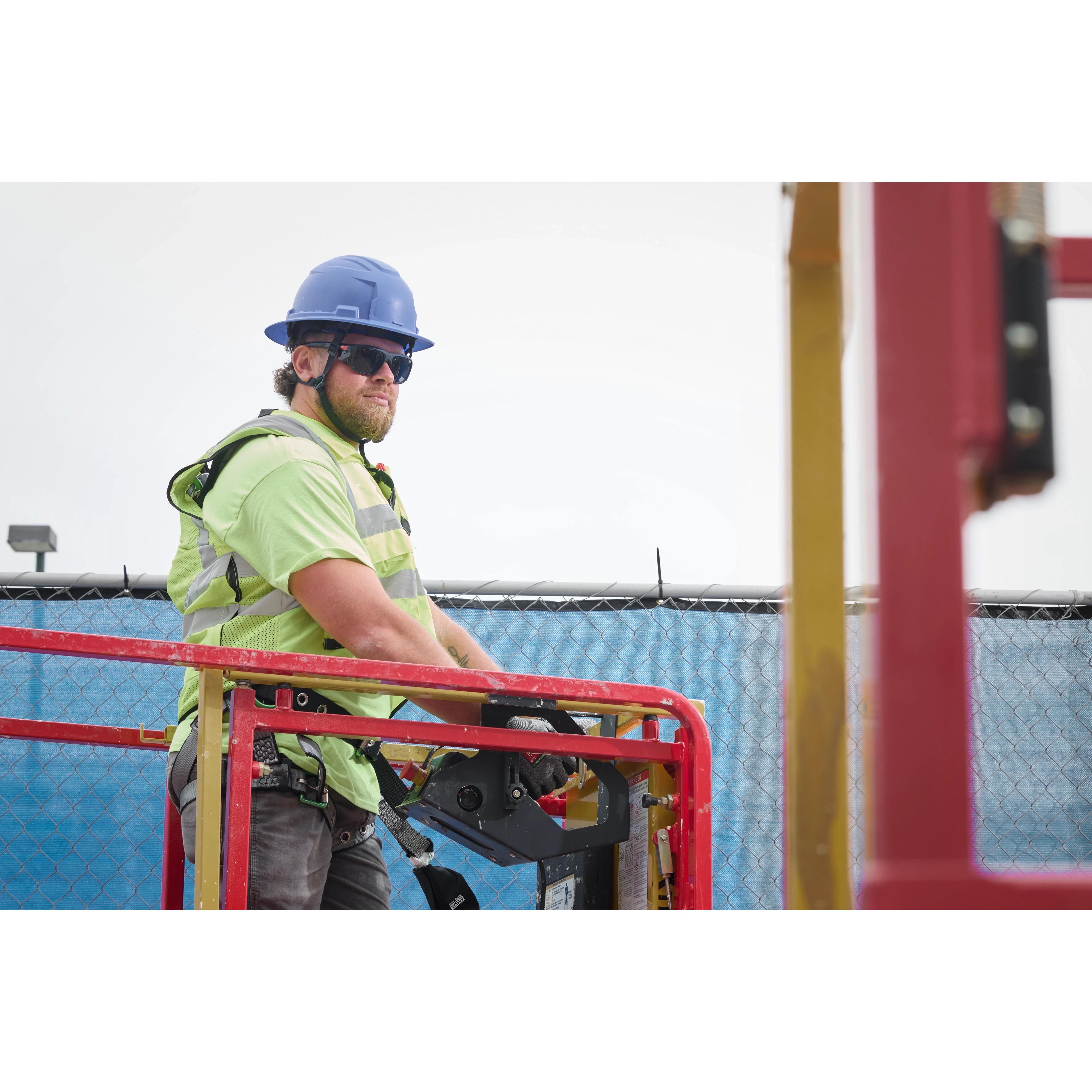 A construction worker in a safety vest, hard hat, and harness stands on a cherry picker. The worker is operating the machine at a worksite. They wear Full Frame Safety Glasses with Removable Side Shields and Tinted Anti-Scratch Lenses. The background features a blue safety net and fence.