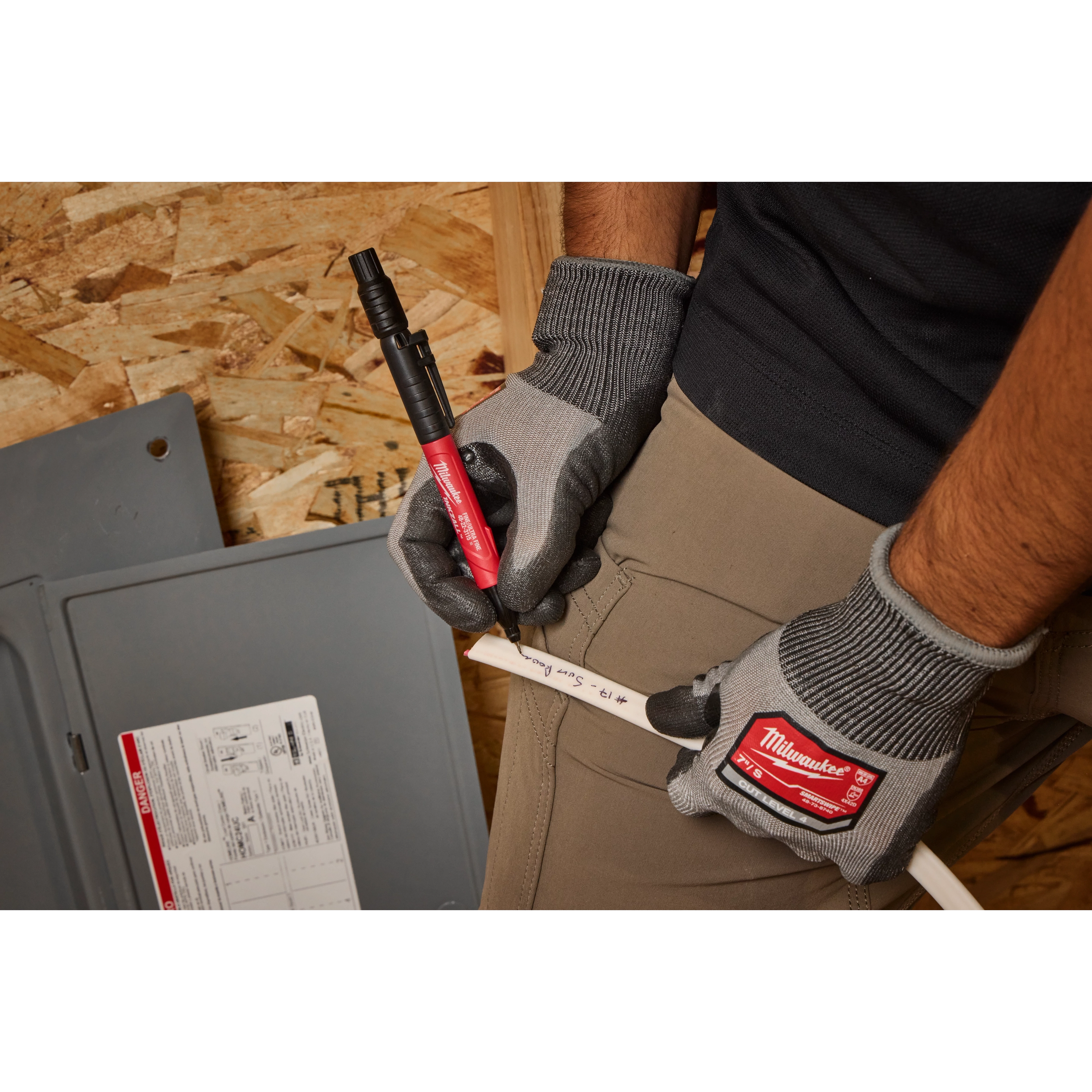 A person wearing gloves is using the INKZALL Double-Sided Black Jobsite Marker—Ultra Fine & Fine Point to write on a white pipe. The black marker, which has a red barrel, is being used in a workshop environment.