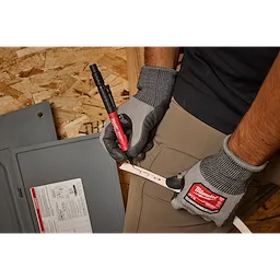 A person wearing gloves is using the INKZALL Double-Sided Black Jobsite Marker—Ultra Fine & Fine Point to write on a white pipe. The black marker, which has a red barrel, is being used in a workshop environment.