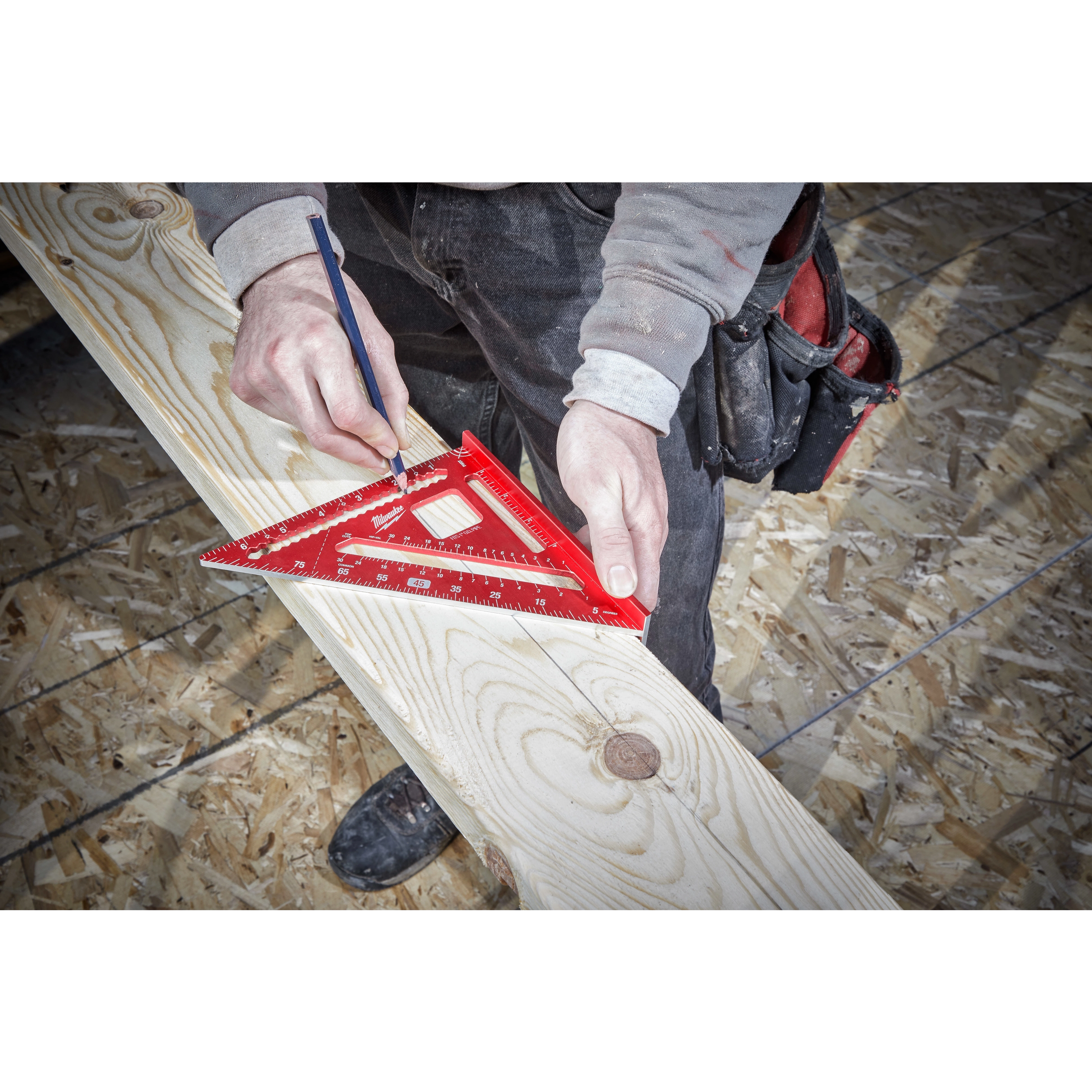 A person marks a wood beam using a red 7 inch Rafter Square and a pencil, with construction materials visible in the background.