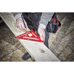 A person marks a wood beam using a red 7 inch Rafter Square and a pencil, with construction materials visible in the background.