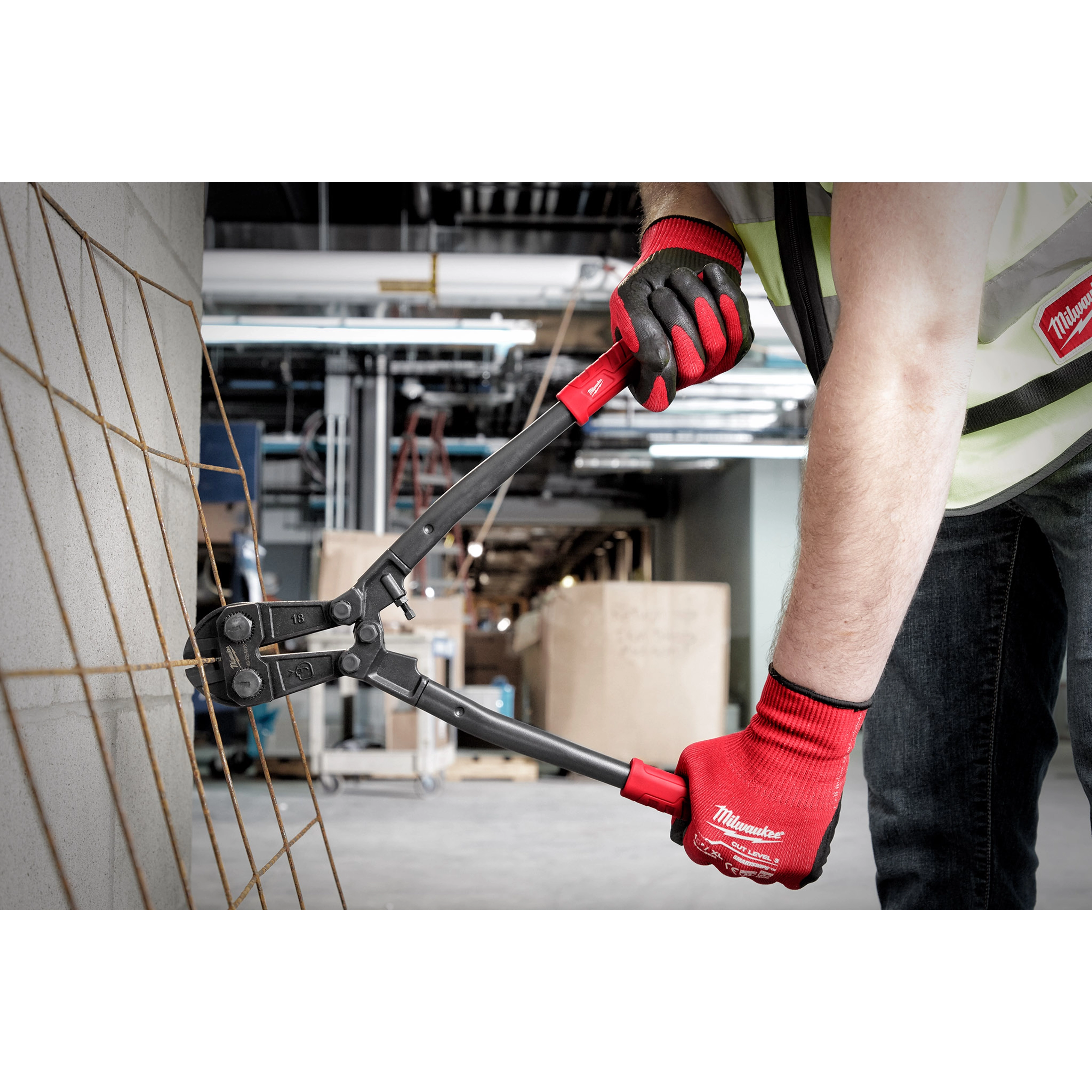 Person using 18" Bolt Cutter with red handles to cut through metal wires at a construction site. They are wearing work gloves and a safety vest.