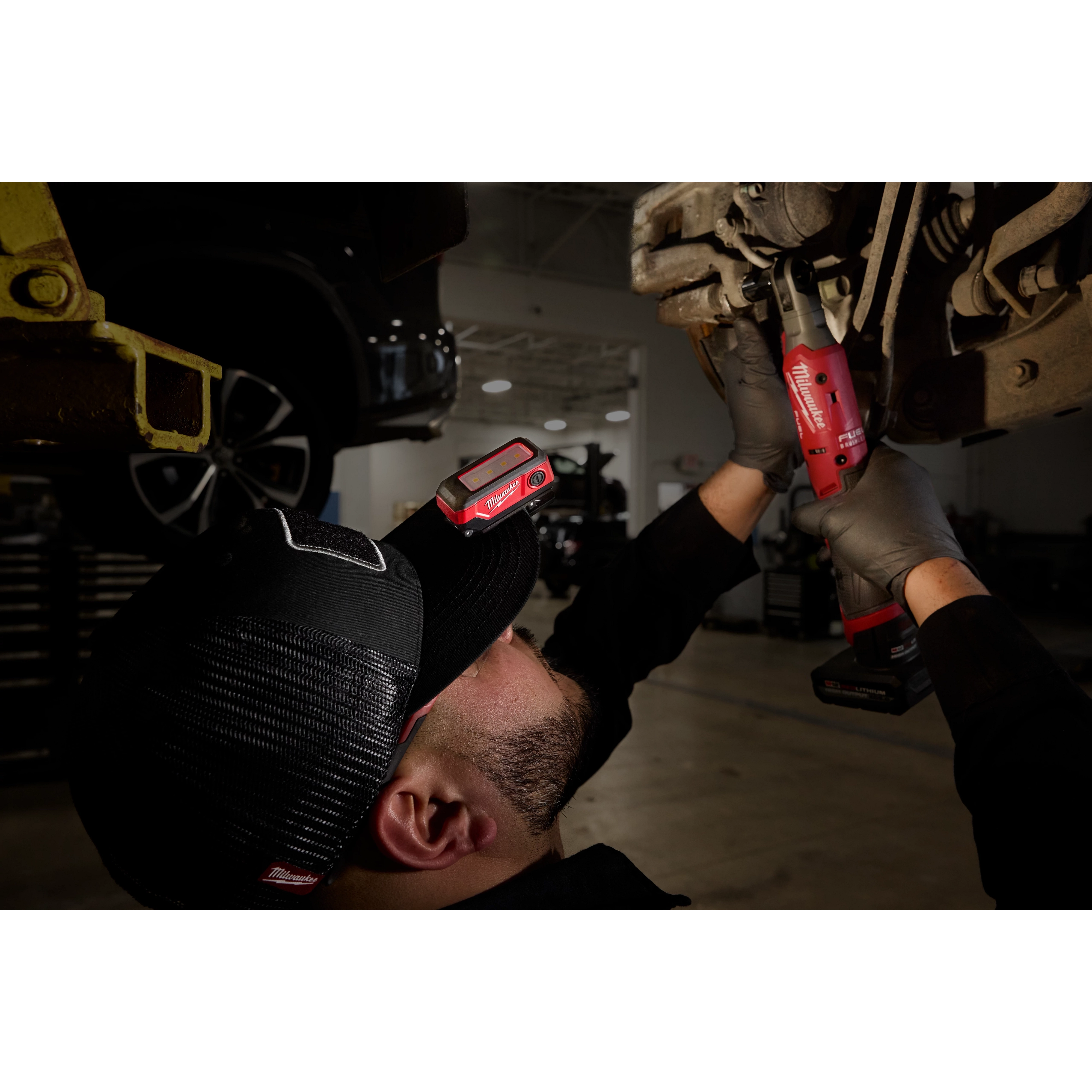 A mechanic works on a vehicle with a Milwaukee Rechargeable Clip Light attached to his cap for illumination.