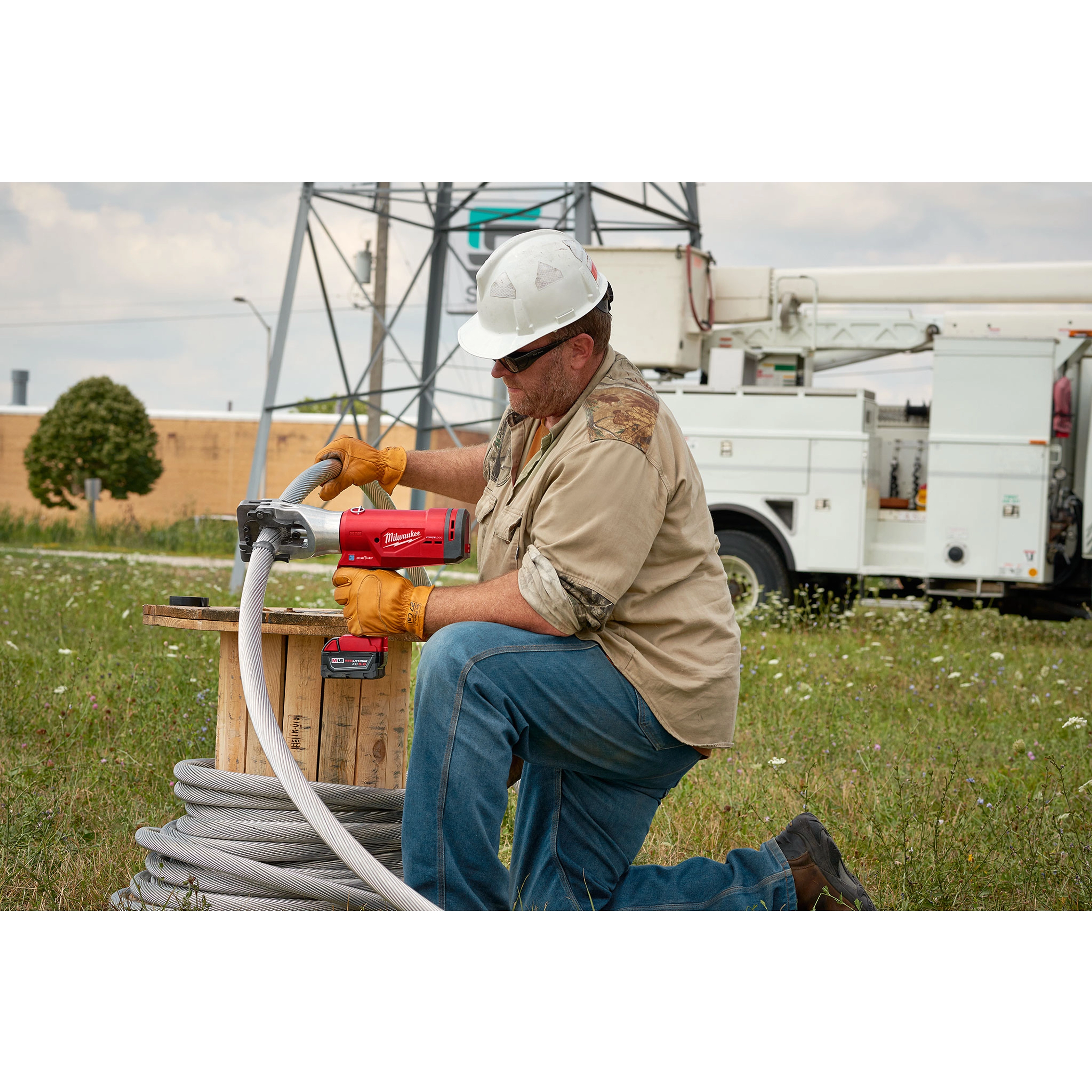A worker uses the FORCELOGIC™ M18™ 1590 ACSR Cutter Kit to cut cables outdoors. The tool is red with ergonomic handles and is connected to a large spool of cable. Industrial equipment and vehicles are visible in the background. The worker is wearing gloves, jeans, and safety gear.