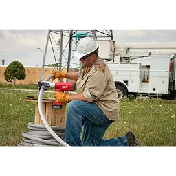 A worker uses the FORCELOGIC™ M18™ 1590 ACSR Cutter Kit to cut cables outdoors. The tool is red with ergonomic handles and is connected to a large spool of cable. Industrial equipment and vehicles are visible in the background. The worker is wearing gloves, jeans, and safety gear.