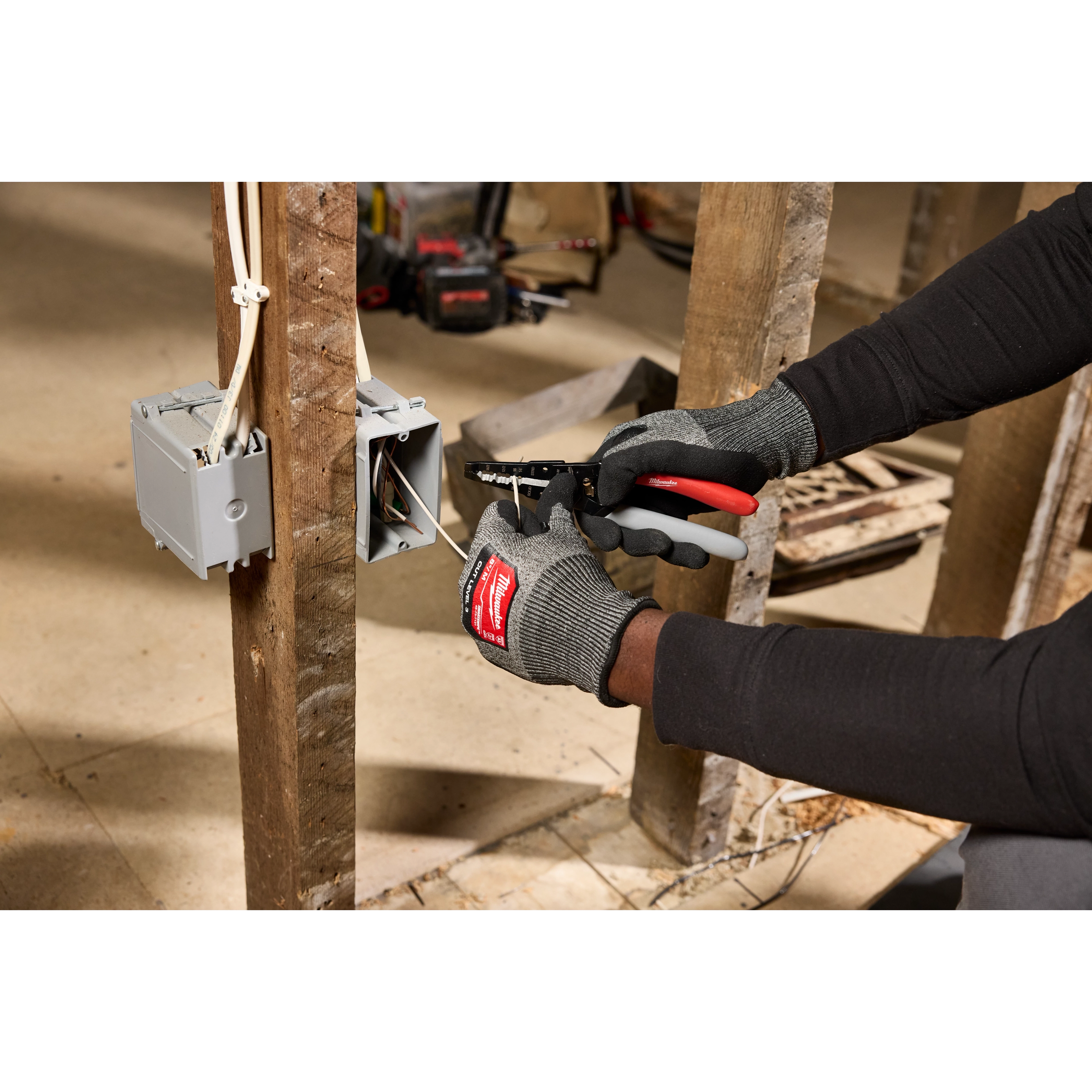 A person wearing gloves uses a 12/2 & 14/2 NM Dipped Grip Wire Stripper & Cutter to strip wires near an electrical box attached to wooden beams.