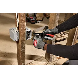 A person wearing gloves uses a 12/2 & 14/2 NM Dipped Grip Wire Stripper & Cutter to strip wires near an electrical box attached to wooden beams.