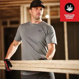 The image shows a man wearing the Hybrid Work Tee - Short Sleeve Feature Shot while holding wooden planks in a construction site.