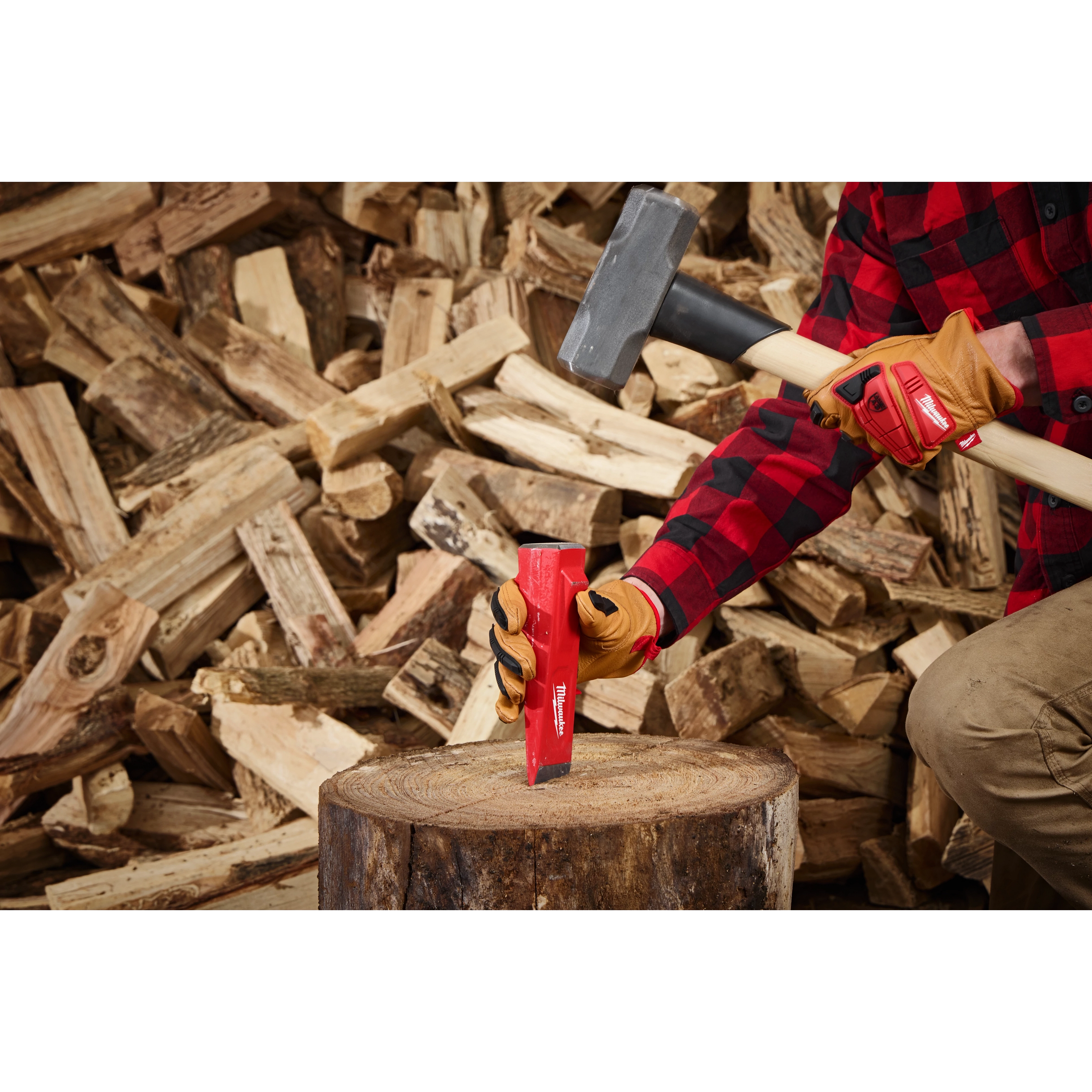 A person wearing gloves and a red plaid shirt holds a mallet and places the 5lb Splitting Wedge onto a tree stump. A large pile of chopped firewood is seen in the background. The wedge is red with white branding, indicating that it is a Milwaukee tool.